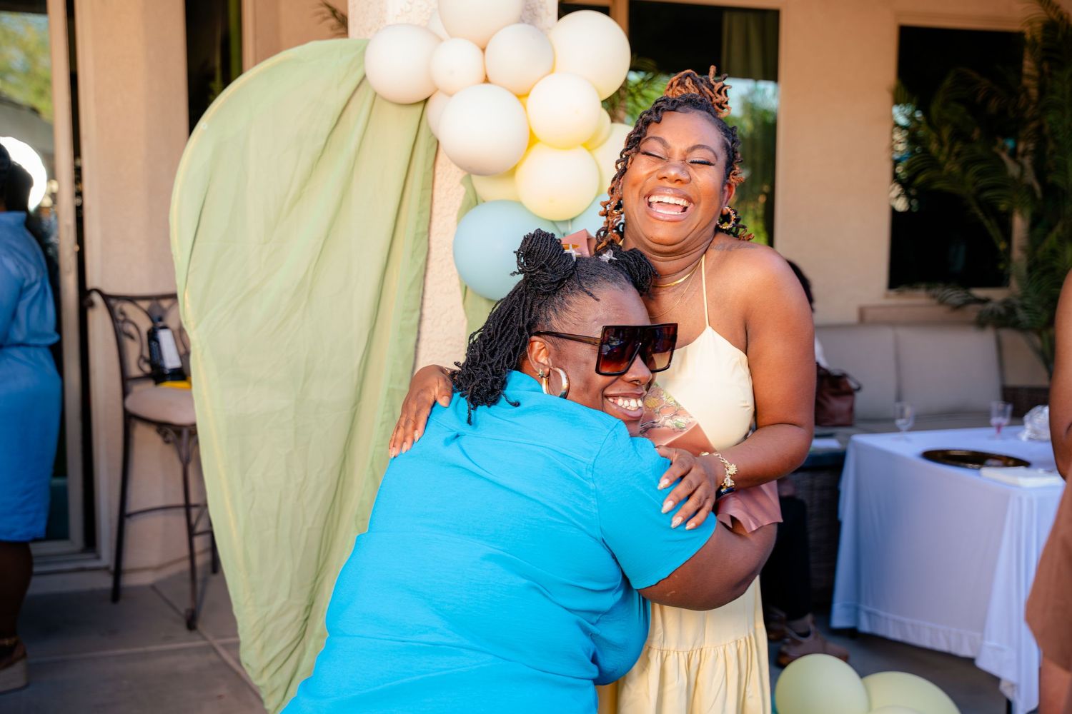 Two women hugging and smiling at an outdoor celebration with balloon arch decor in green, white, and yellow.