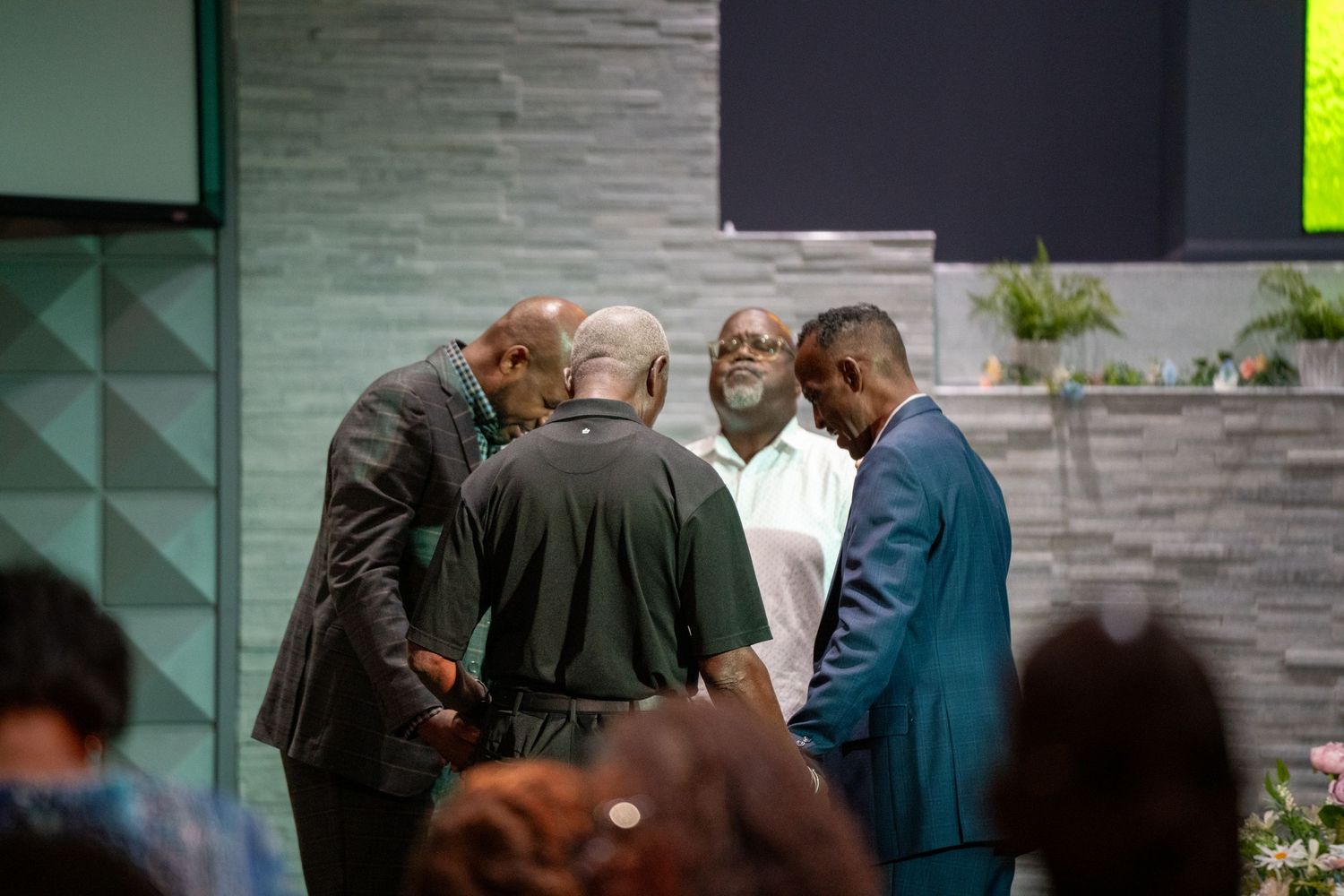 A group gathers in prayer at a modern church with textured walls and ambient lighting.