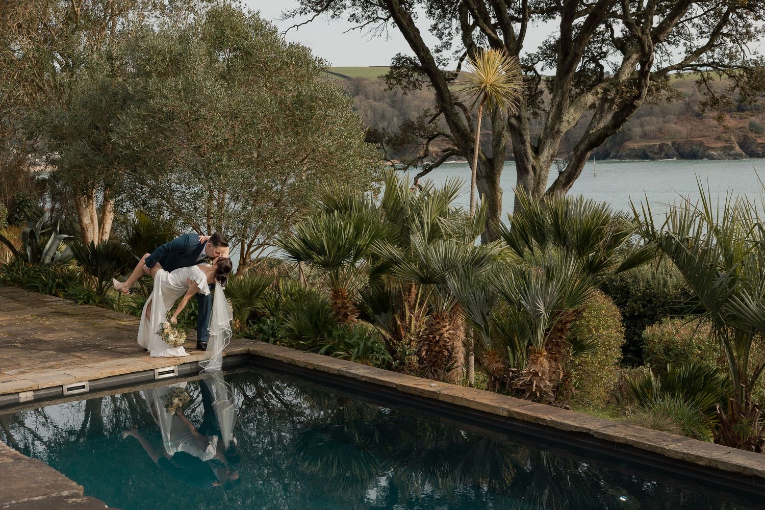 Bride and Groom kissing next to swimming pool at The Moult in Salcombe with the sea behind them