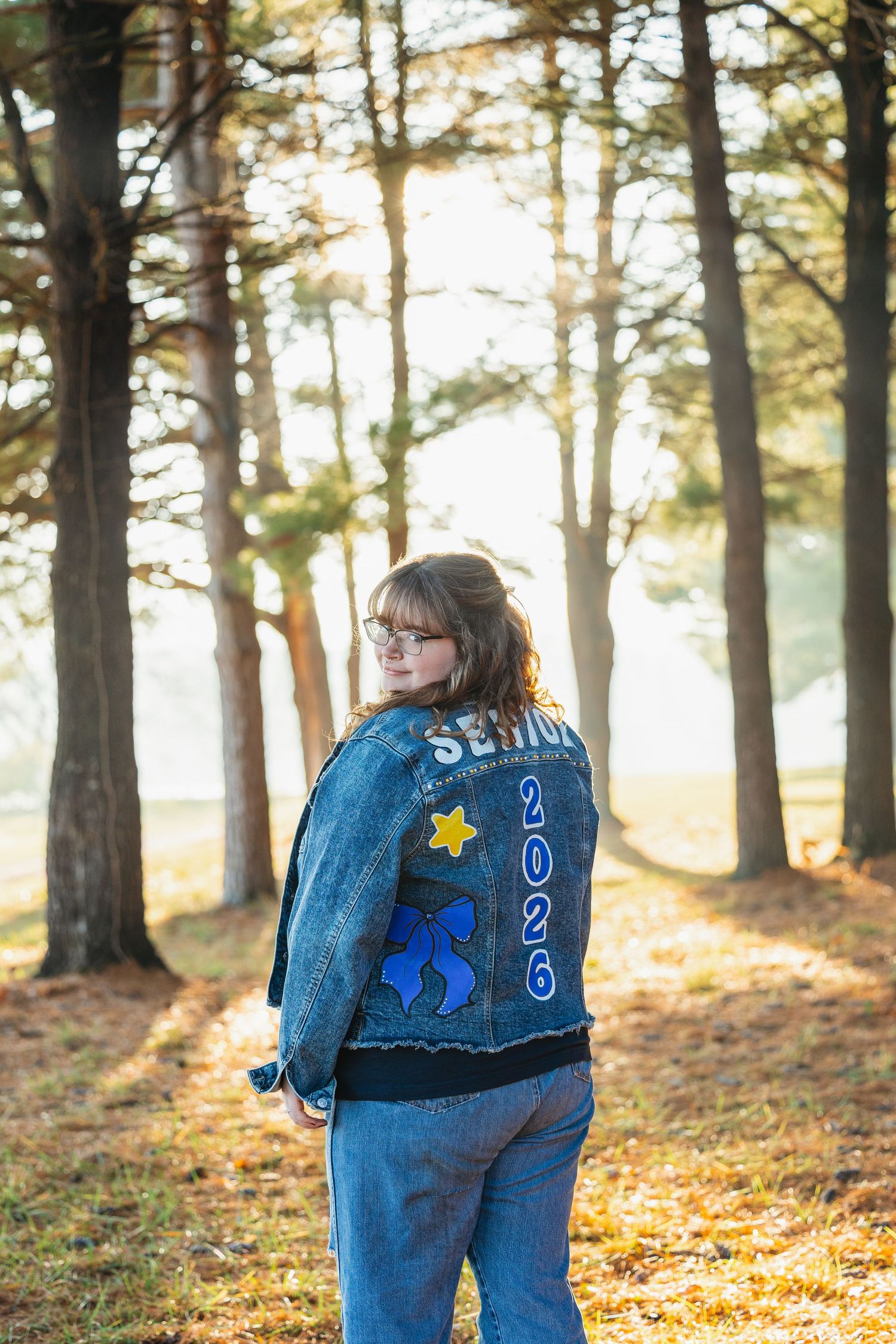 Person wearing denim jacket stands among tall pine trees in autumn forest with golden sunlight filtering through.