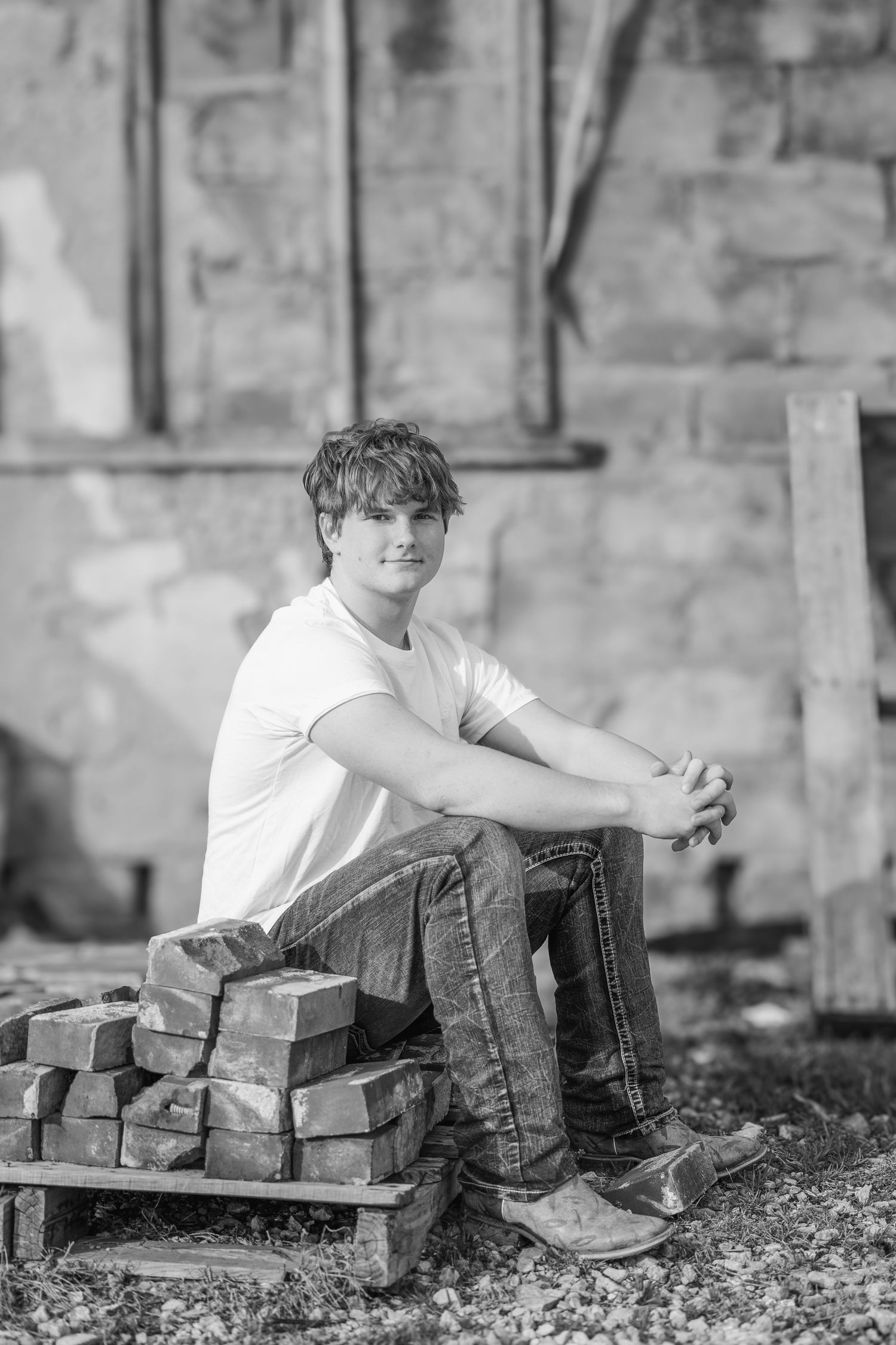 A vintage black and white photograph showing a person sitting on bricks in a casual pose against a stone wall.