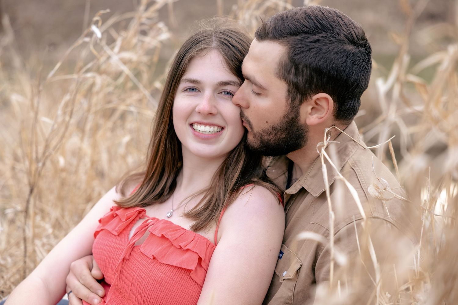 Intimate engagement photos of couple in coral dress and casual attire amid tall grass.