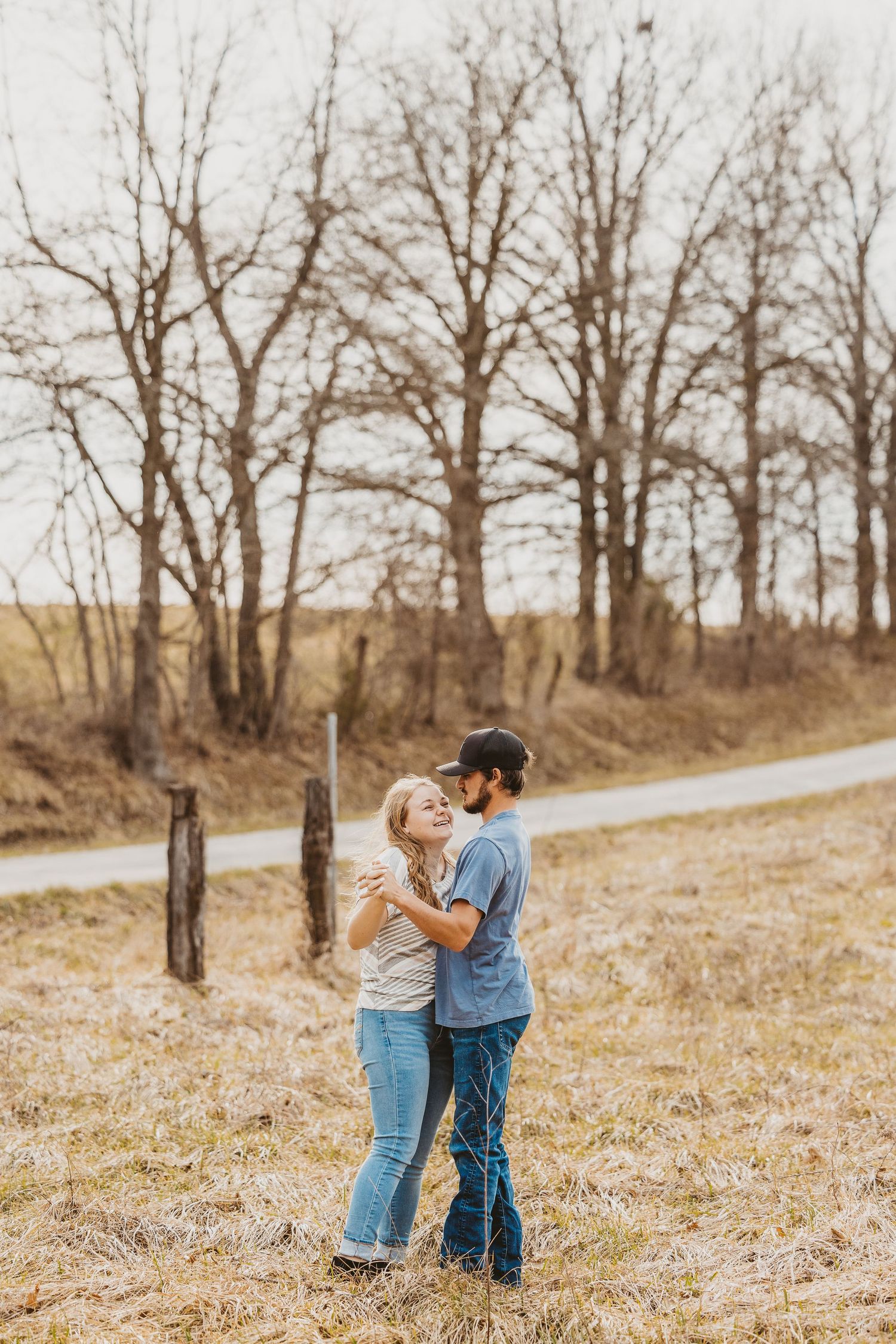 A couple embraces and laughs together in a rural field with bare trees and a dirt path in the background.