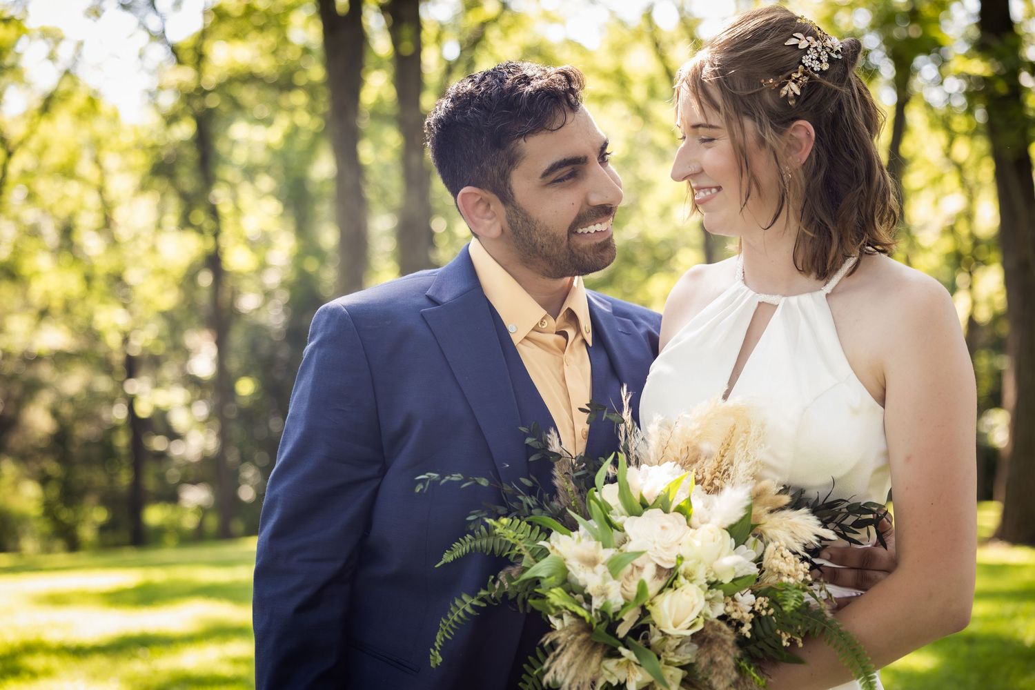 A couple shares an intimate moment in a sunlit garden setting while holding a white floral bouquet.