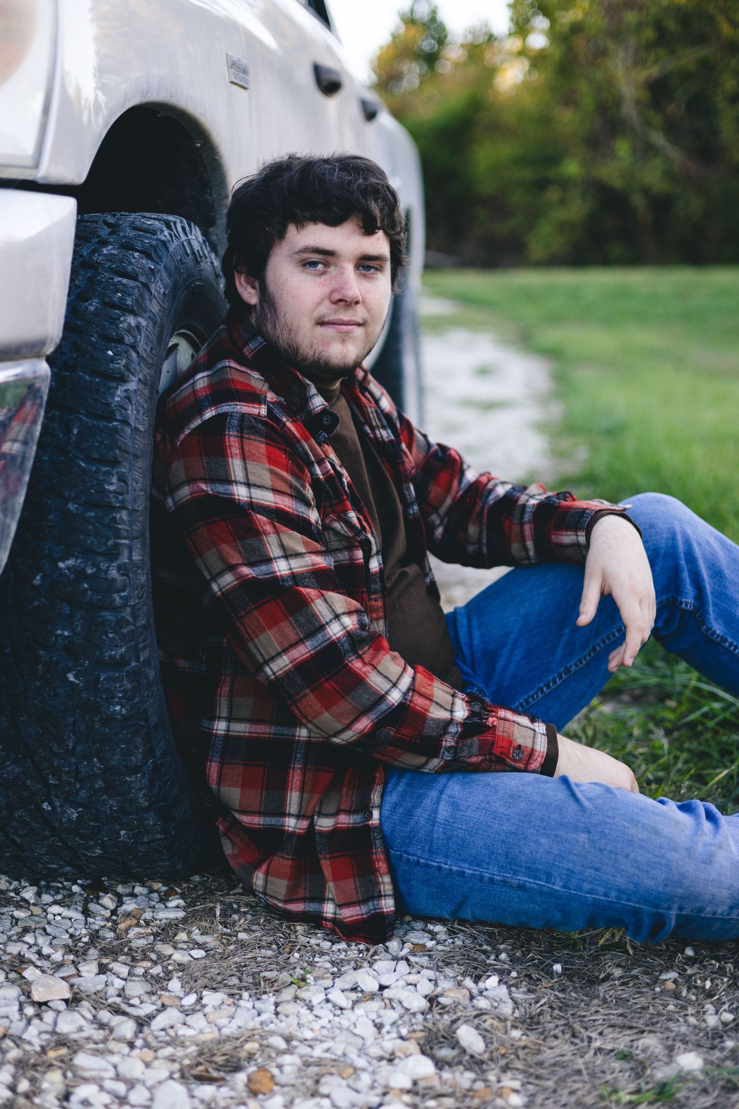 Person in plaid shirt and jeans relaxing against white truck tire on gravel.