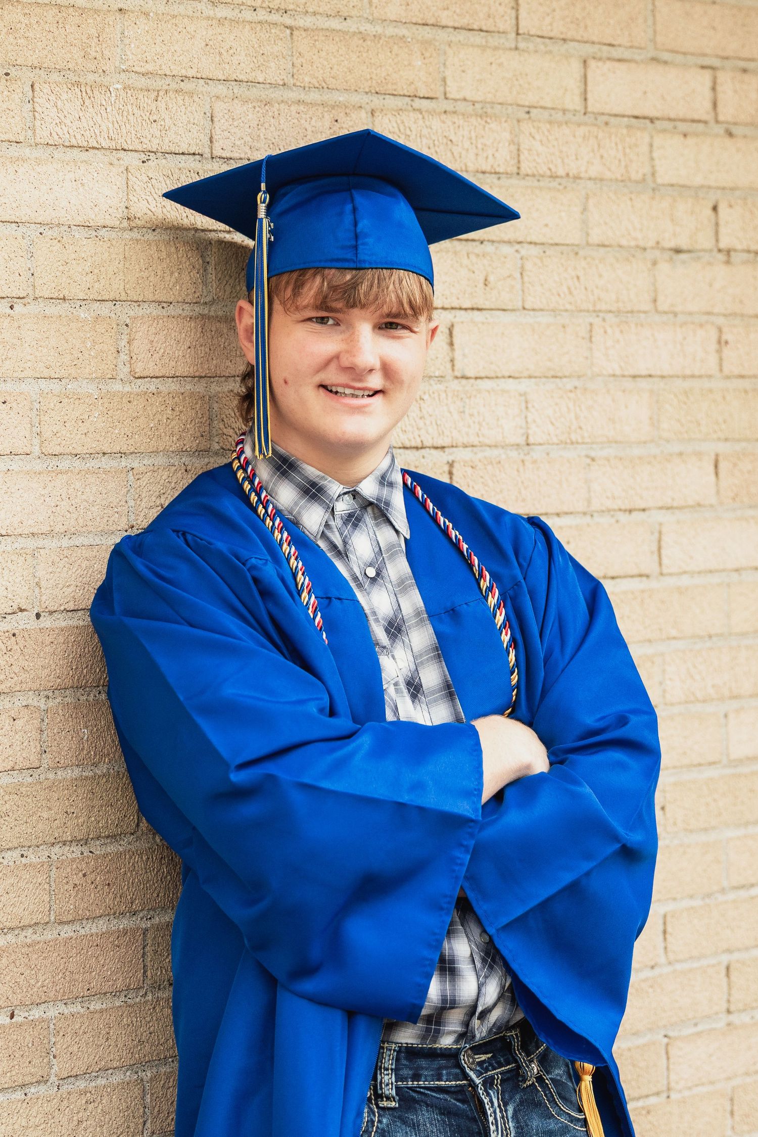 Smiling young man in blue graduation cap and gown with honor cords, arms crossed, leaning against brick wall.