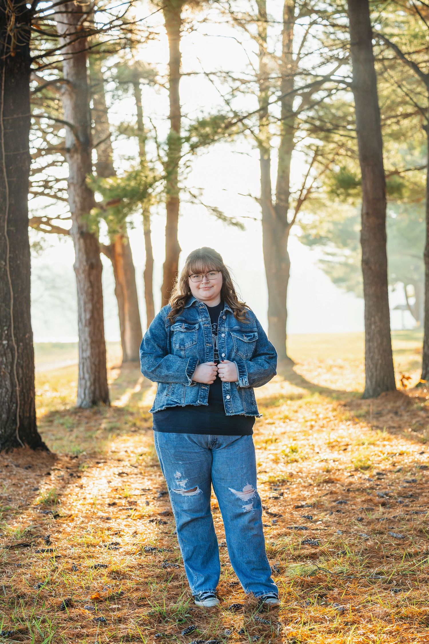 Toddler in denim overalls plays among tall pine trees on an autumn day with golden sunlight filtering through.
