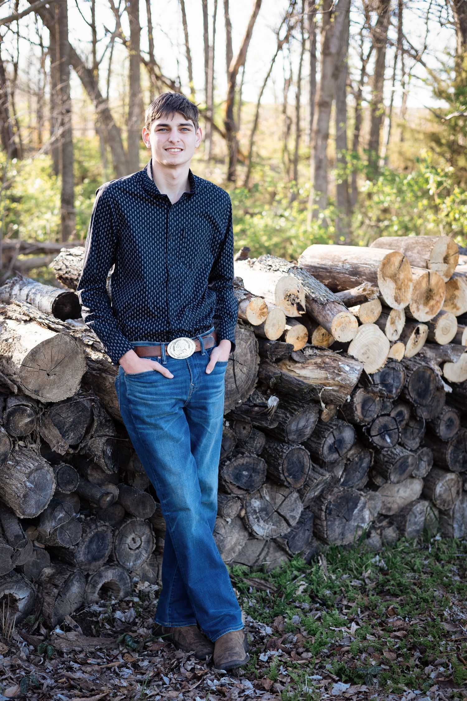 Person in polka dot shirt leaning against stacked firewood in a wooded outdoor setting.