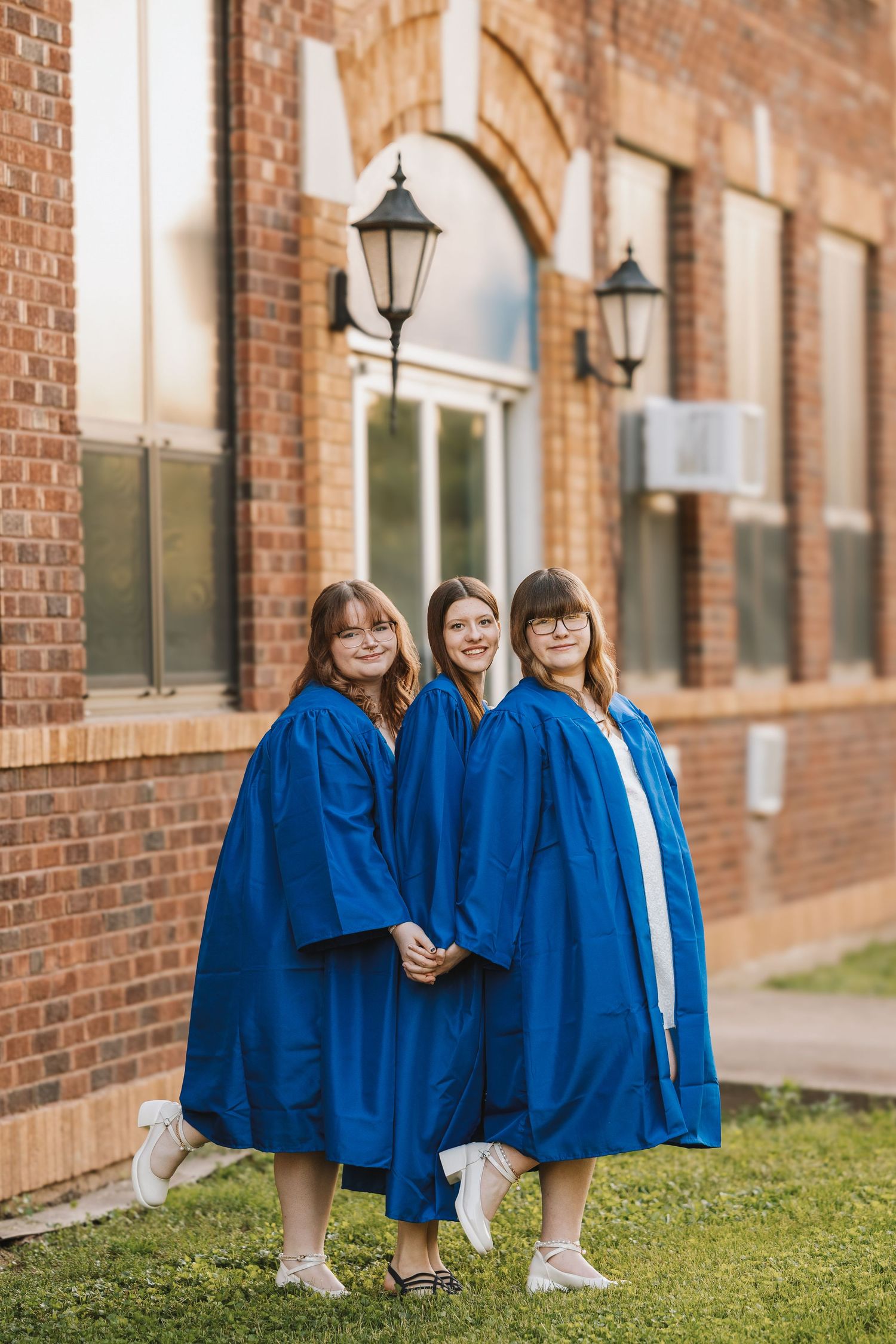 Three girls in blue graduation gowns pose together outside a brick building.