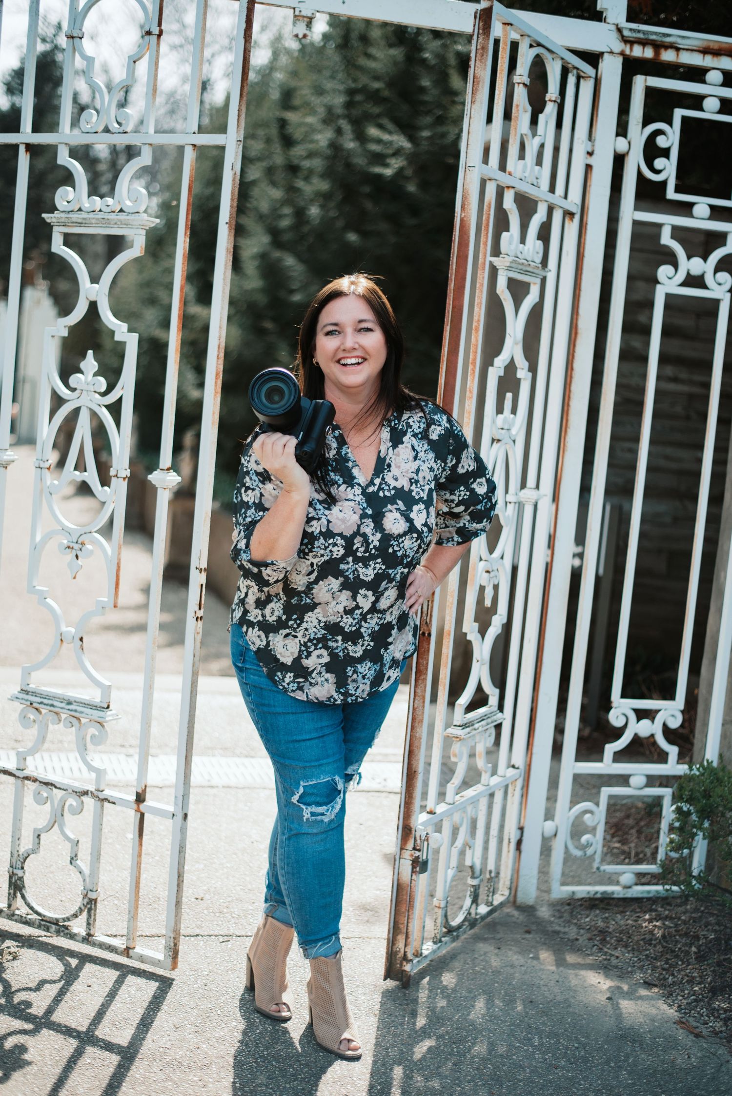 Smiling woman holding a camera poses by ornate white iron gates in a floral top and jeans.