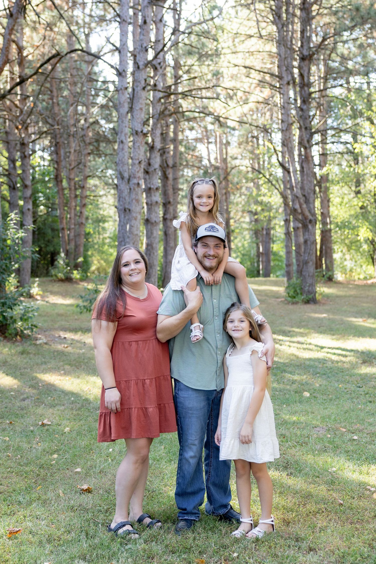 A family poses together in a sunlit forest setting wearing casual summer outfits.