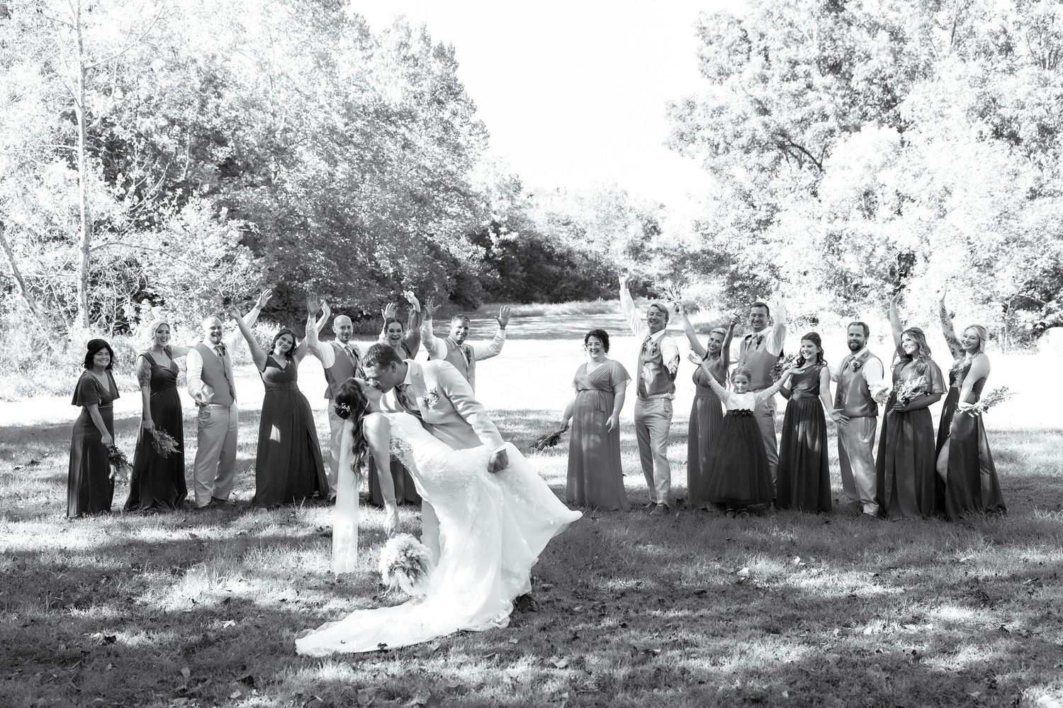 A wedding party poses in a natural outdoor setting with trees and greenery in the background.