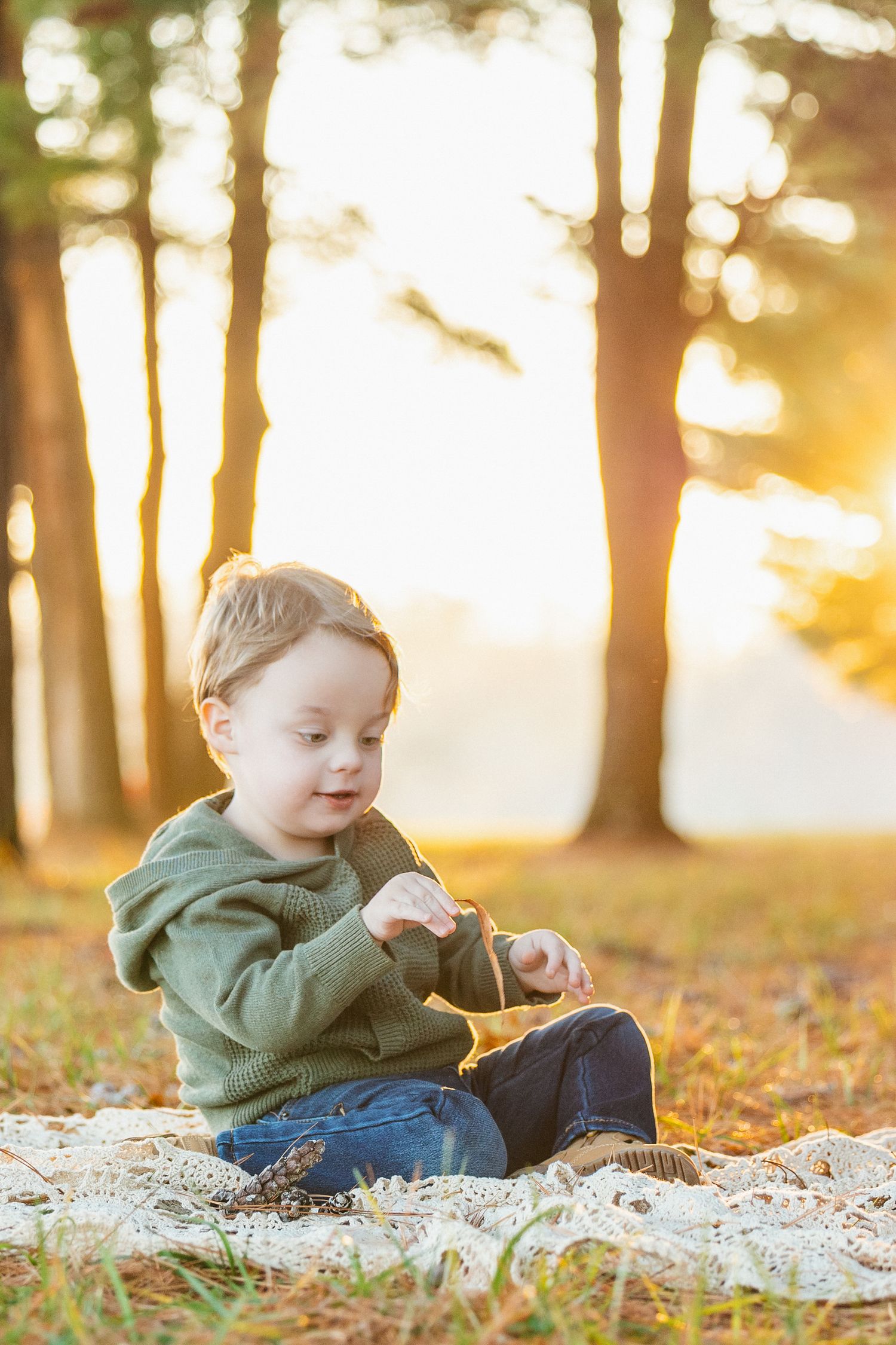 A child in a green hoodie plays in autumn leaves during golden hour sunset in a wooded park setting.