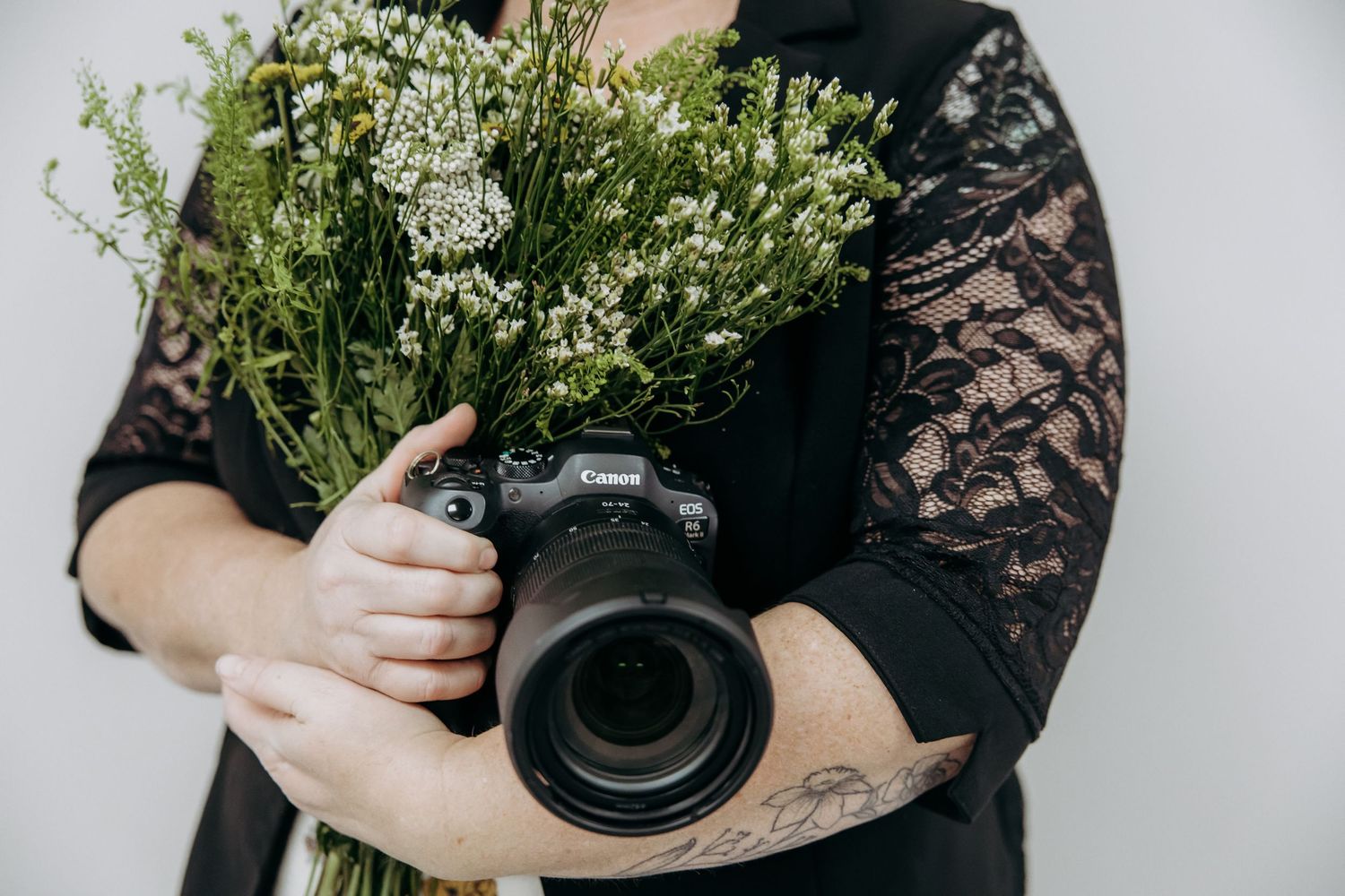 A photographer with tattoos holds a professional camera against a backdrop of green foliage and white wall.