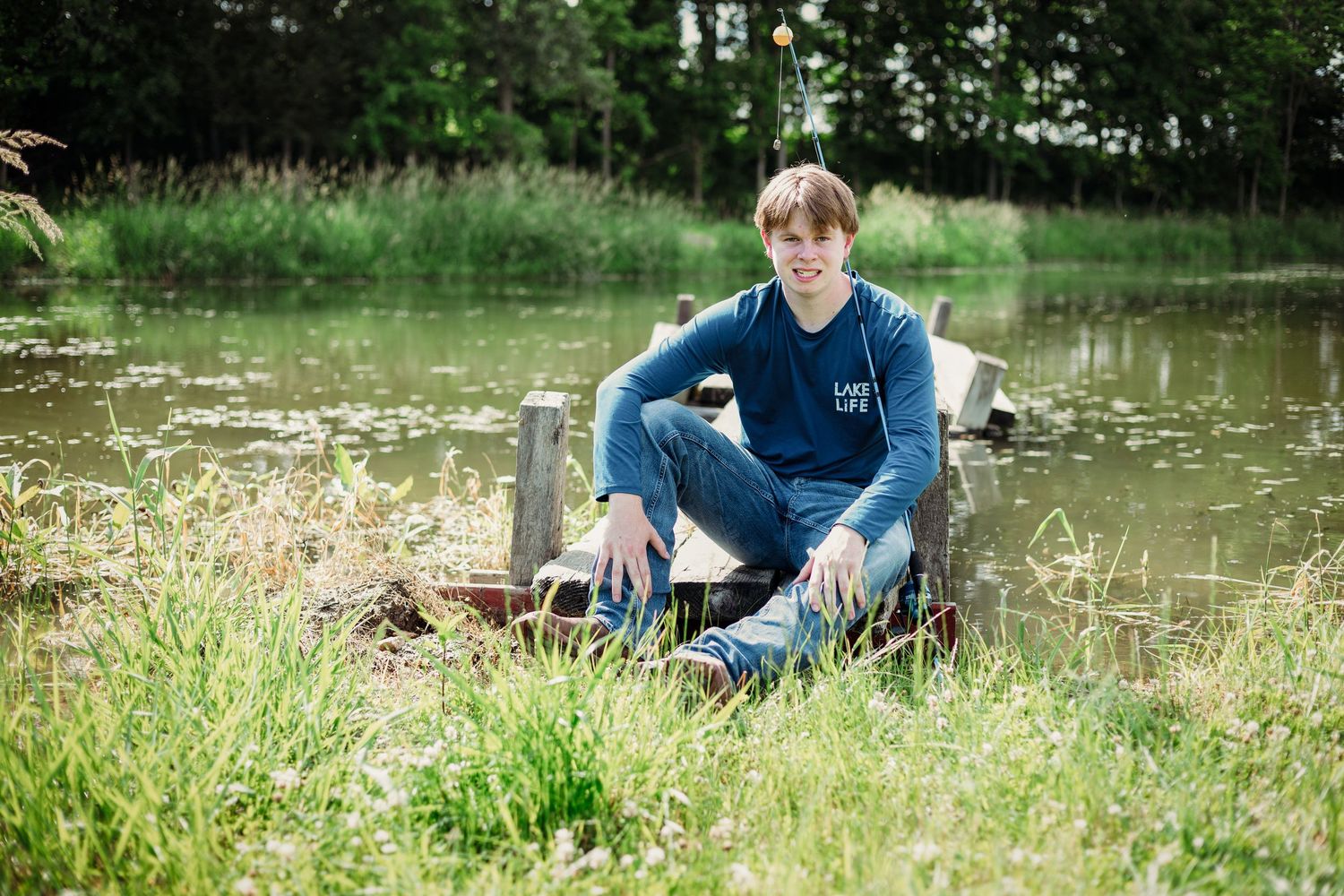 Person sitting casually by a serene pond surrounded by lush grass and natural scenery.