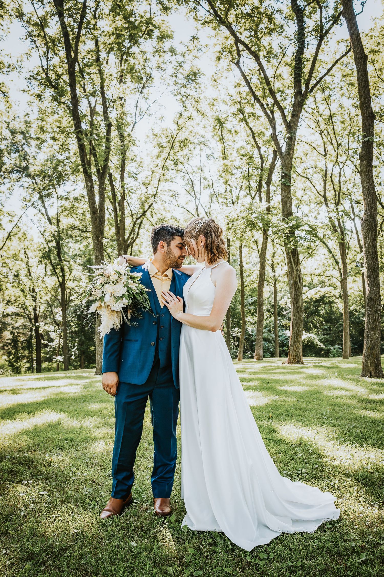 Bride and groom share a romantic moment in a lush green park, bride in white gown, groom in blue suit holding bouquet.