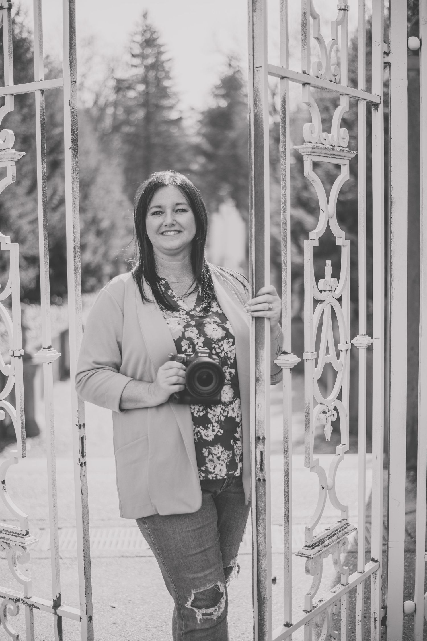 A photographer stands between ornate metal gates holding a camera in a black and white portrait.