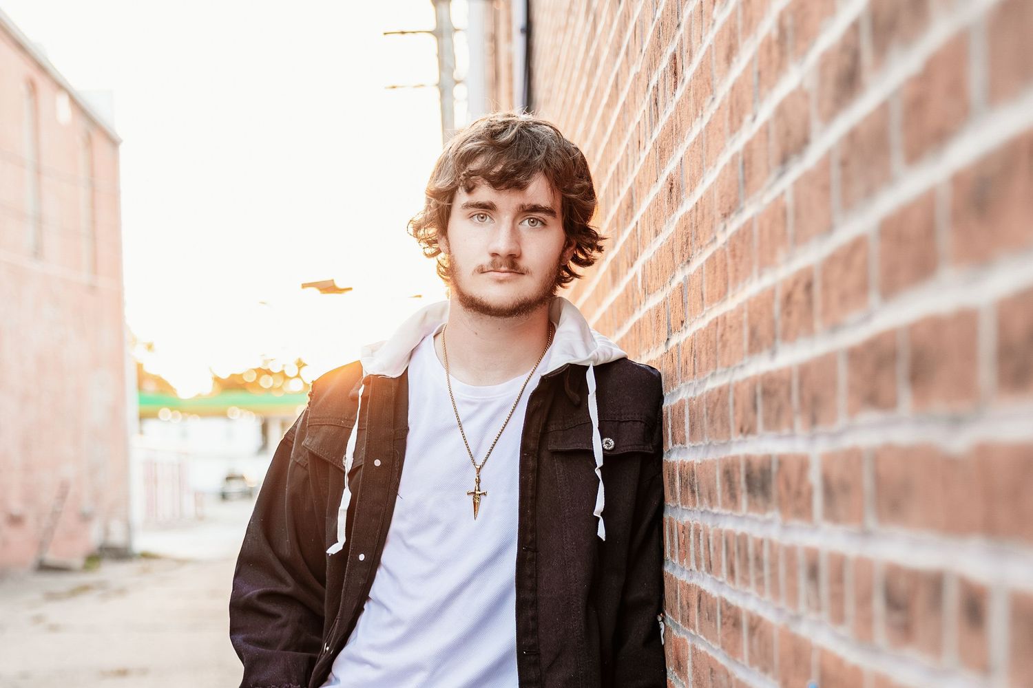 A person in a brown jacket leans against a brick wall in an outdoor setting.