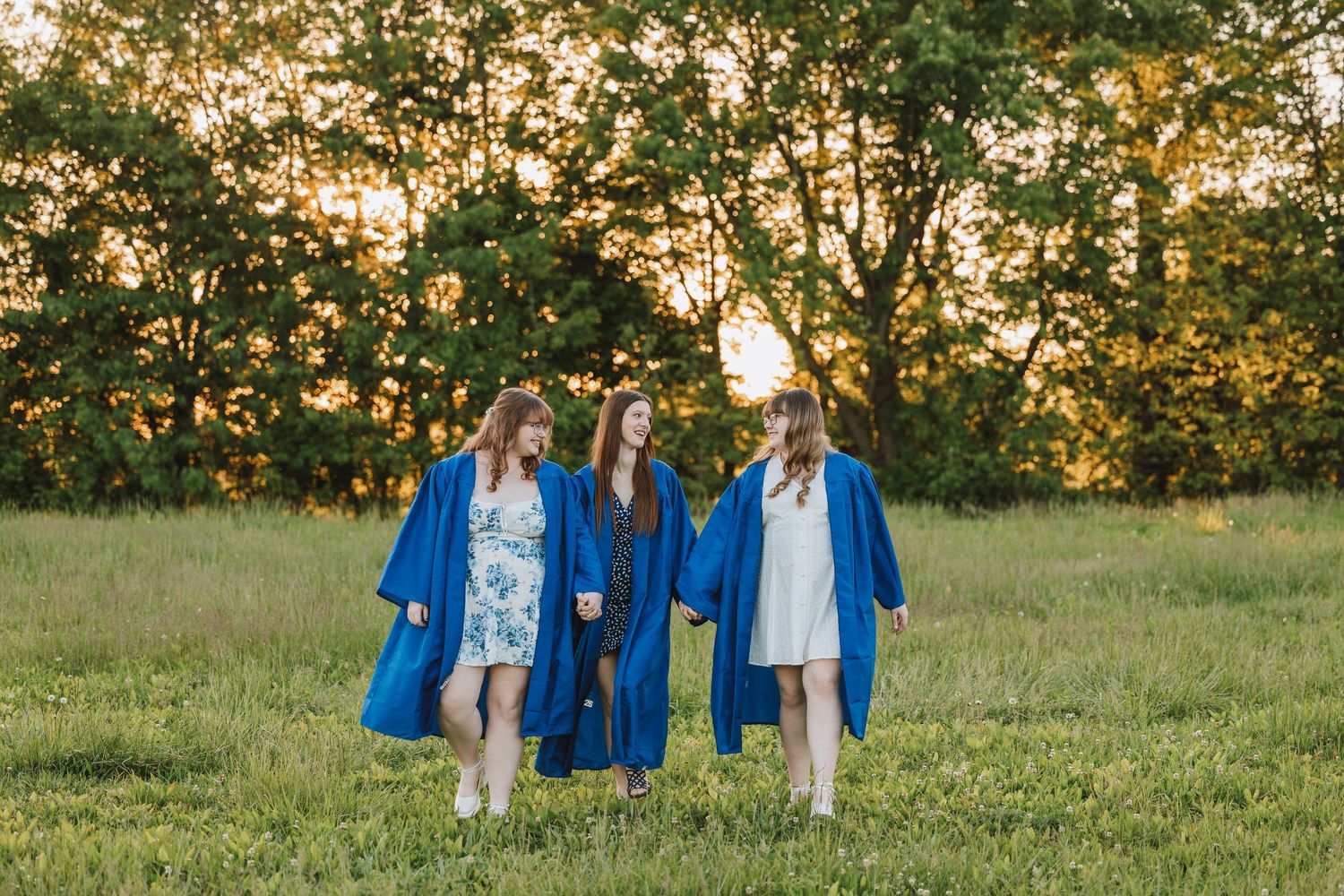 Three young women in blue graduation gowns walking through a green field at sunset.