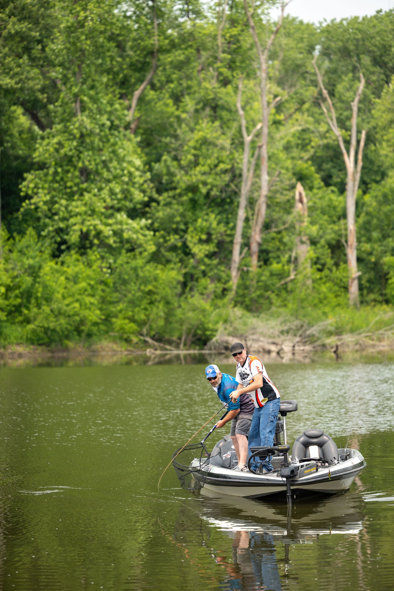 Two people fishing from a small motorboat on a tranquil lake surrounded by lush green trees.