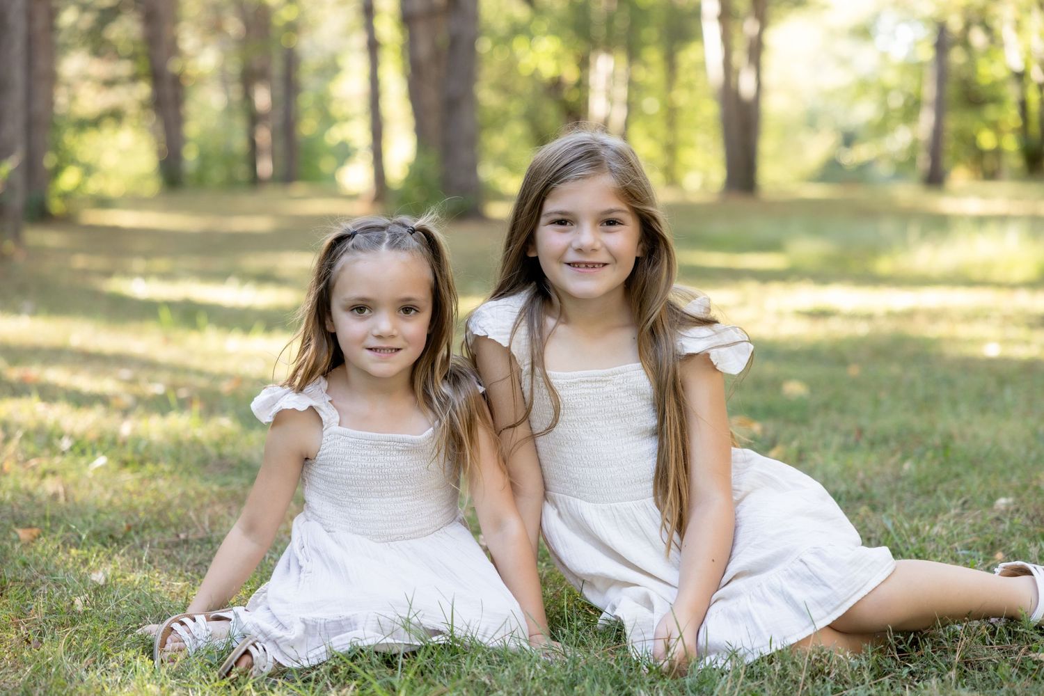 Two children in white dresses sit together on grass in a sunlit wooded area during summer.