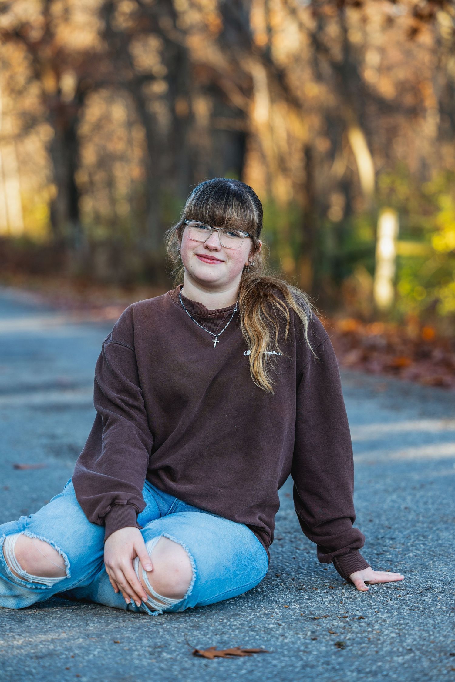 A person in a brown sweatshirt and jeans sits casually on a paved path during autumn.