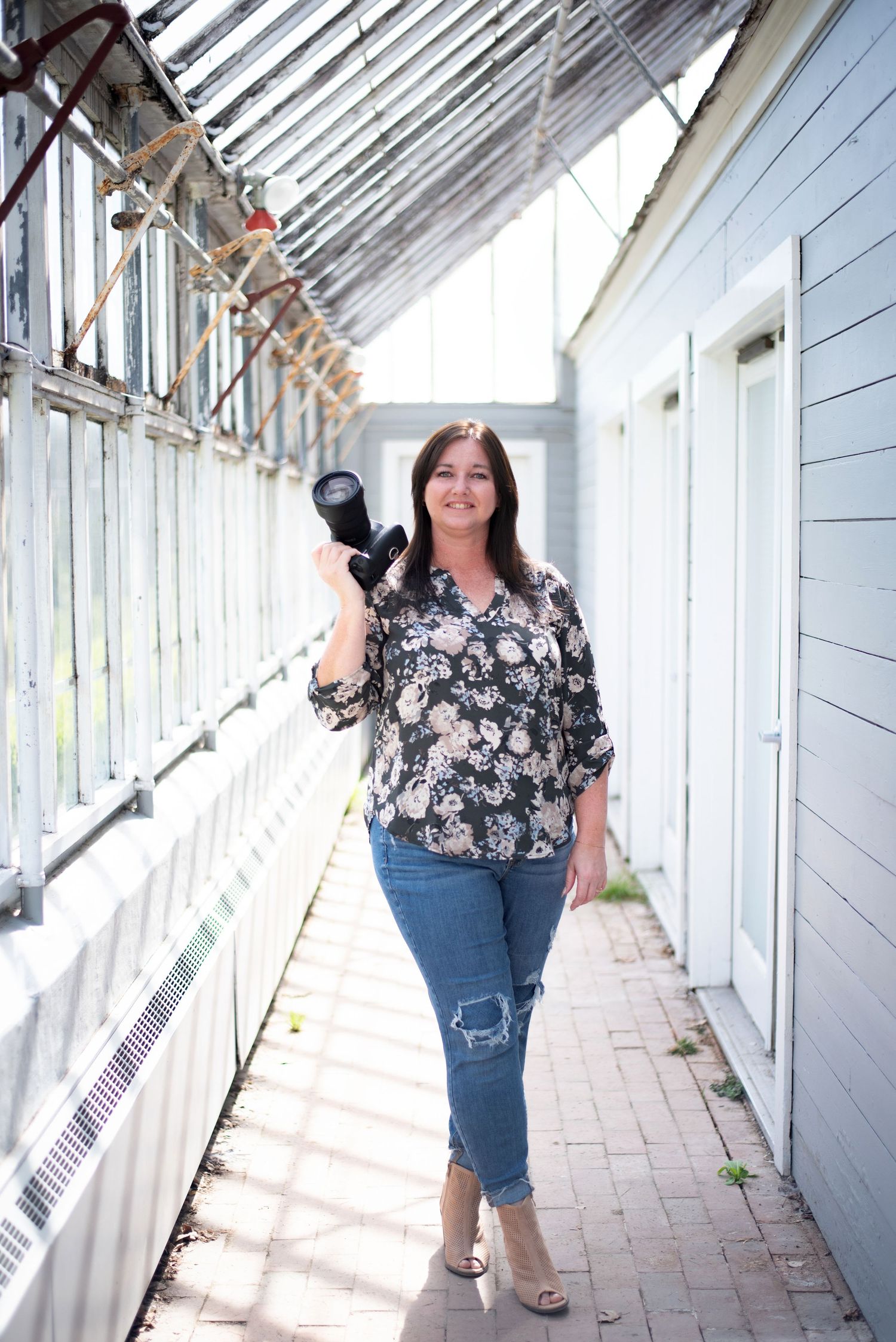 Professional photographer with camera standing in bright sunlit greenhouse corridor wearing floral top and jeans.