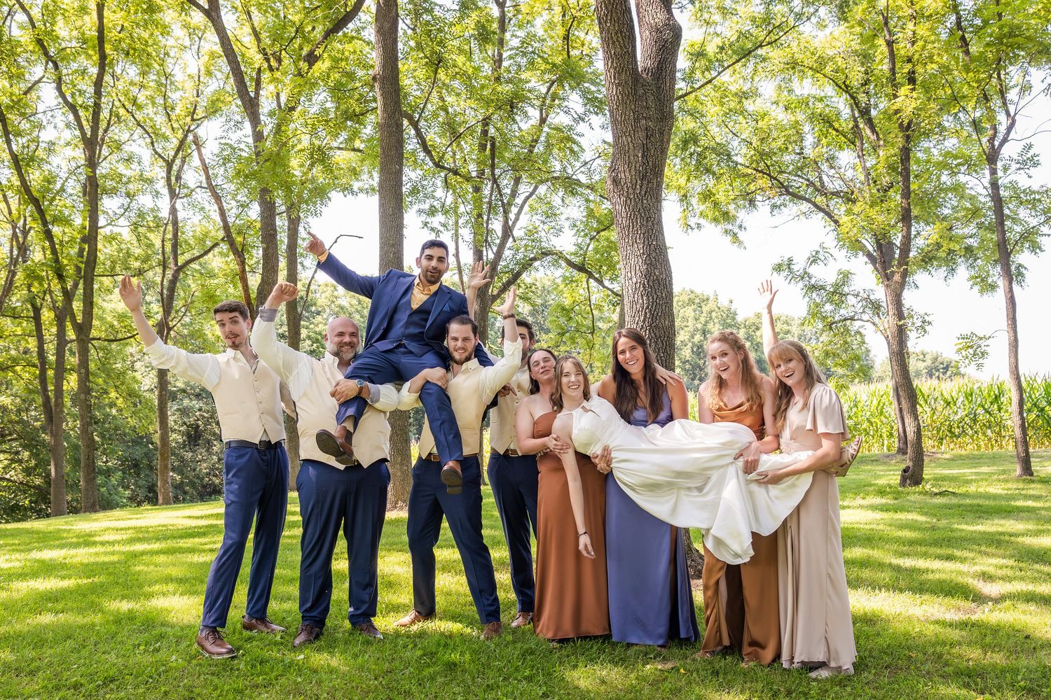Large wedding party posing for outdoor portraits under green trees.