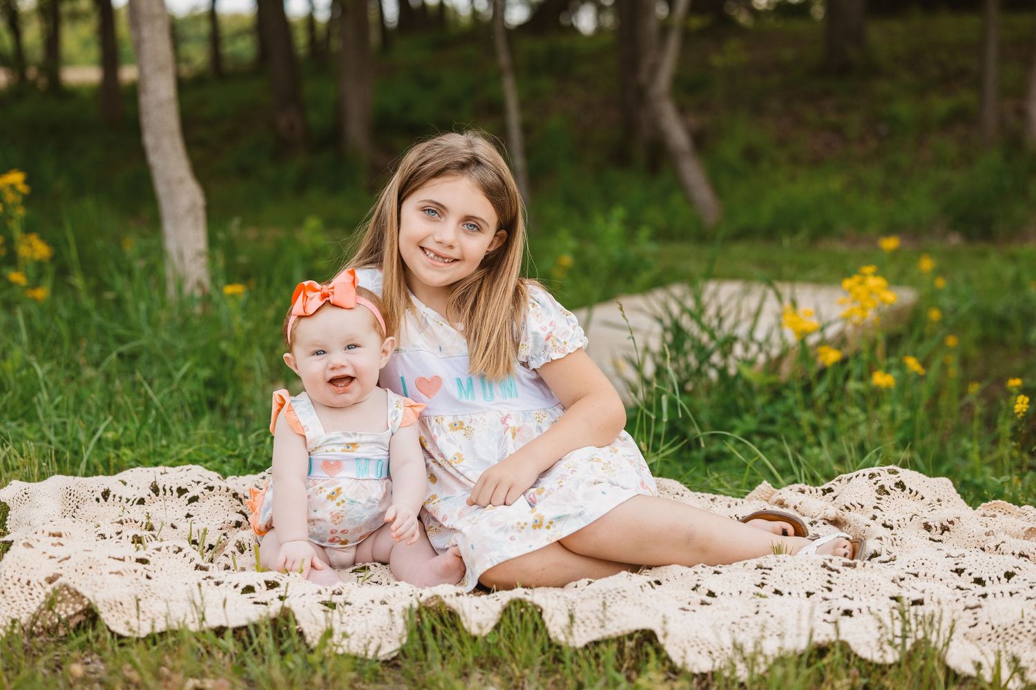 Two sisters sitting on a lace blanket outdoors, older girl smiling and hugging a laughing baby in a sunny green park.