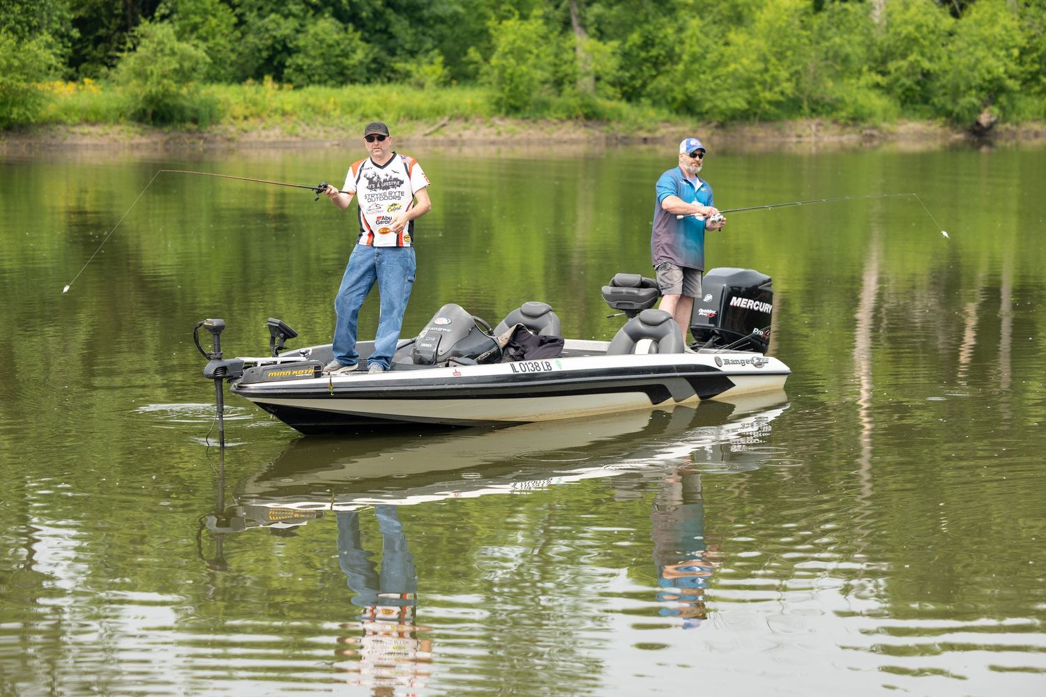 Multiple fishing boats spread out across a calm lake surrounded by green trees during a summer day.