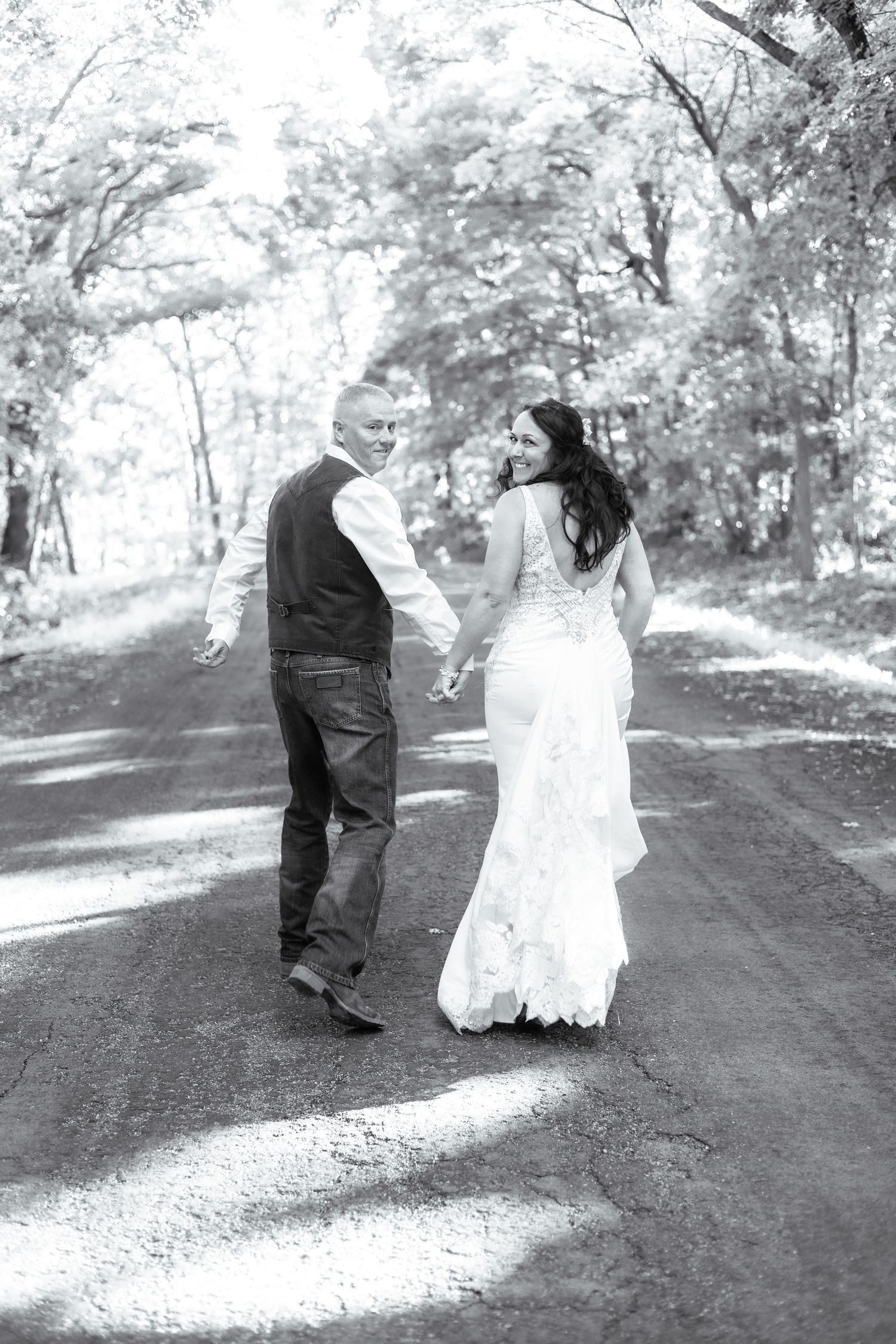 Black and white photo of a couple walking down a tree-lined path in wedding attire.