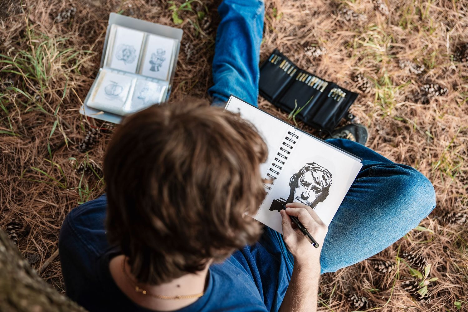 Overhead view of someone sketching a portrait in a notebook while sitting on brown grass with art supplies nearby.