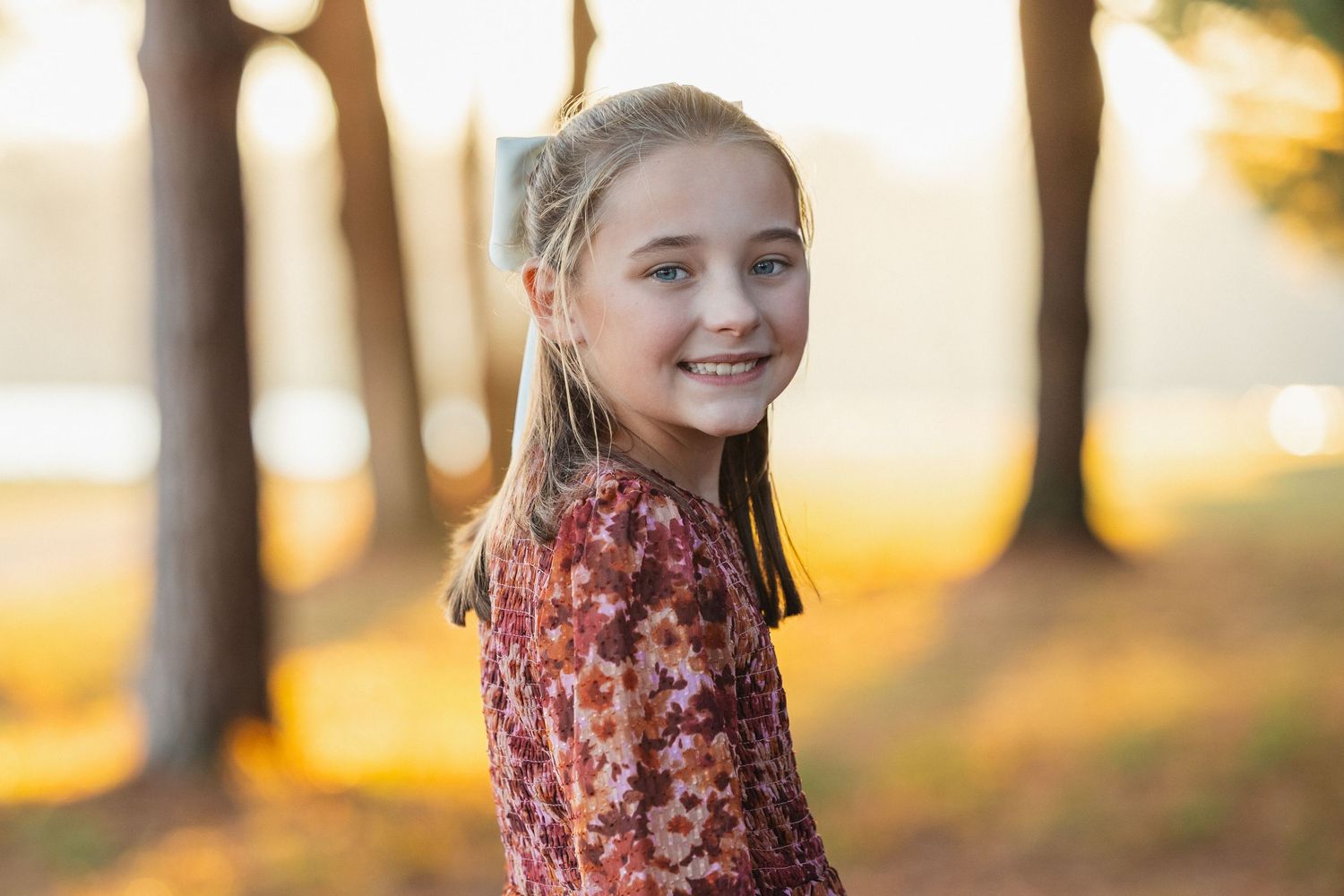 Young person in a floral print dress smiling outdoors during golden hour with sunlit trees in background.