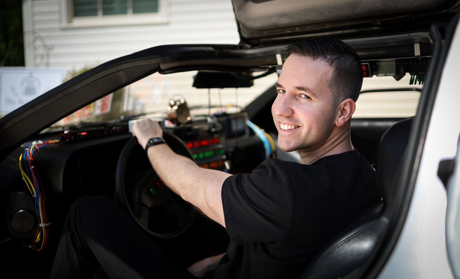 A smiling man sits in the driver seat of a white sports car, wearing a black t-shirt.