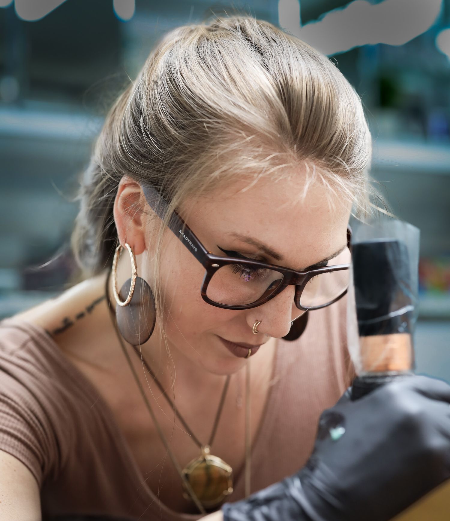 Woman with glasses and hoop earrings concentrating while tattooing, wearing black gloves in a professional studio.