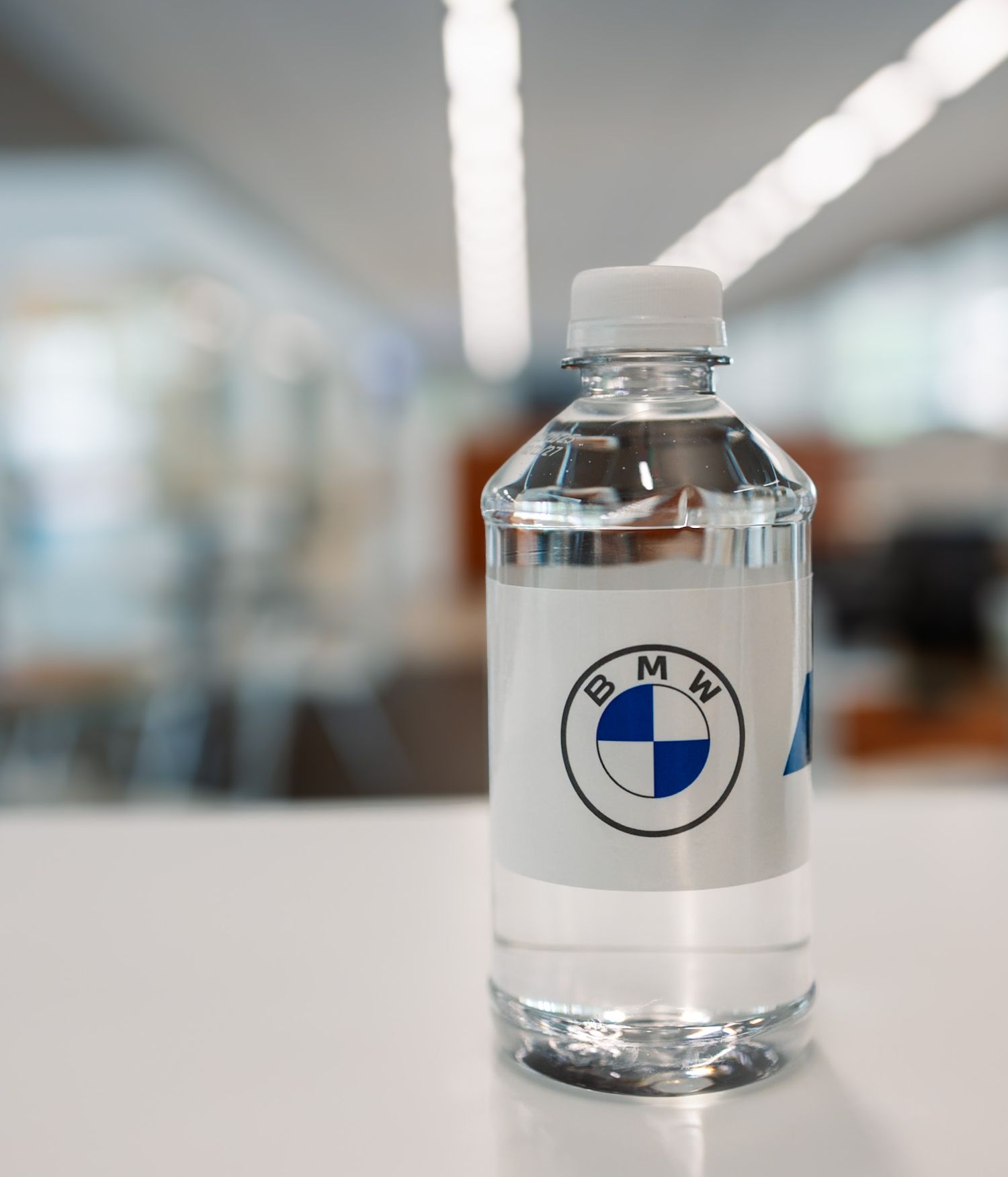 Clear plastic water bottle with BMW logo label on white table in blurred office background.