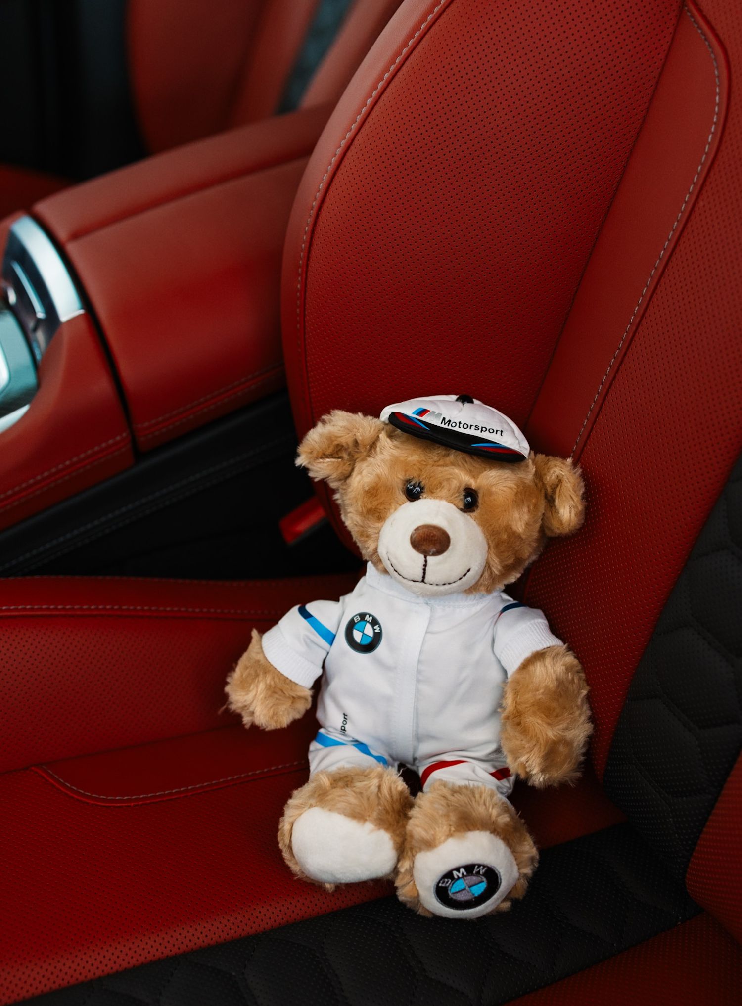 BMW Motorsport teddy bear in white racing suit sitting on red leather car seat.