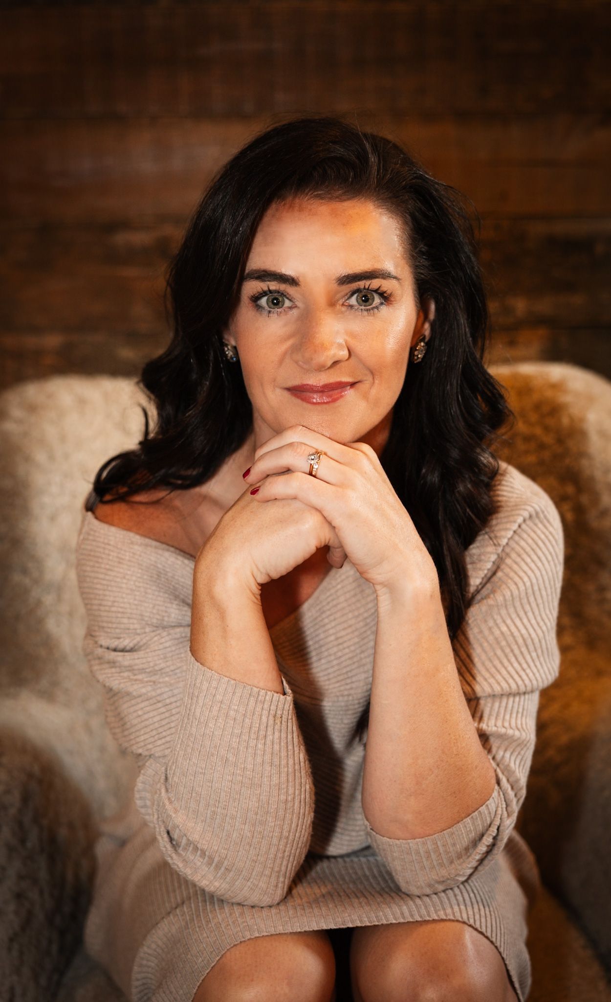 A smiling woman with dark hair in a beige off-shoulder sweater poses with hands clasped, seated in a cozy chair.