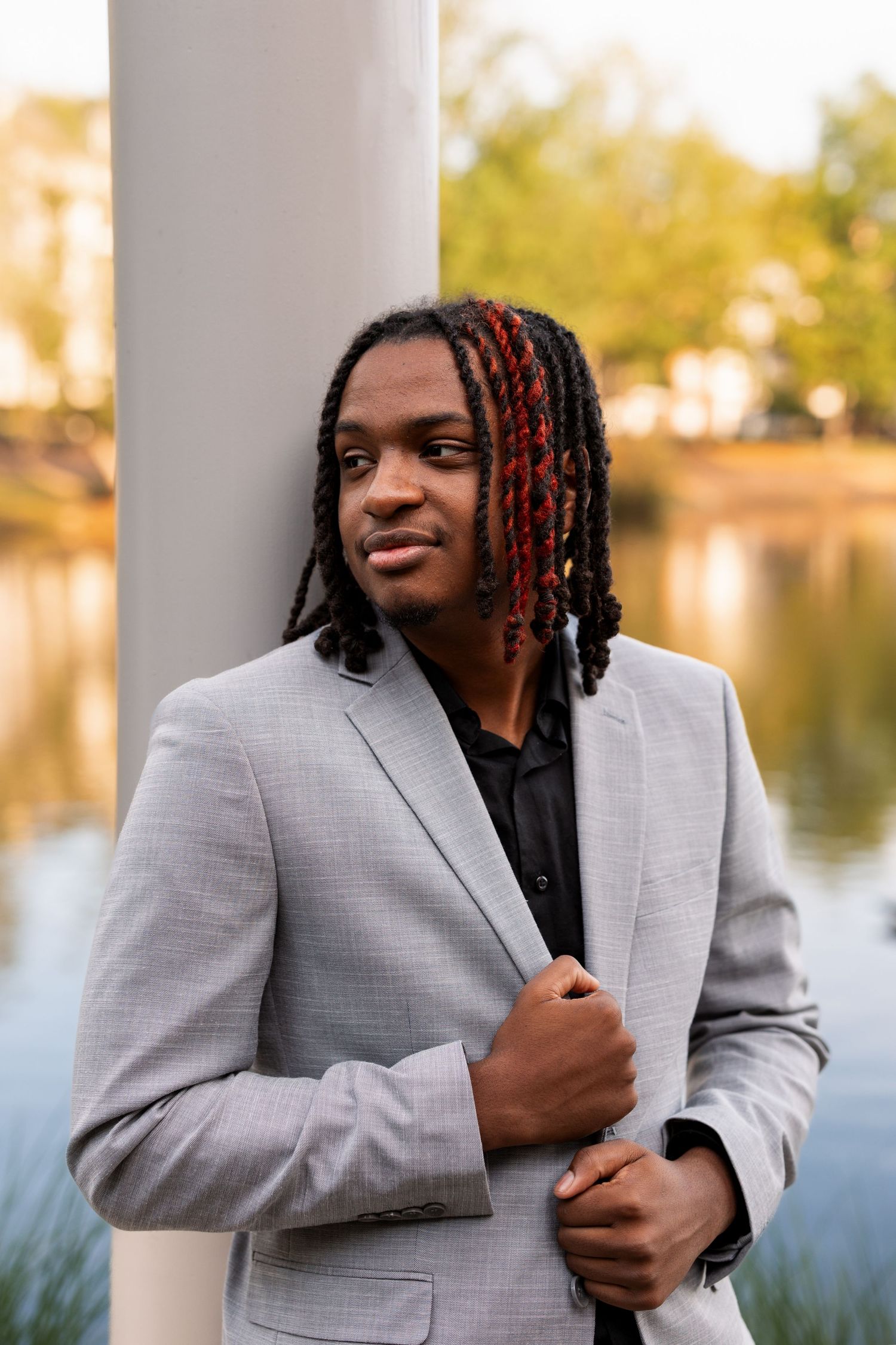 Young Black man with red-tipped dreadlocks wearing a gray blazer poses confidently near a column by a serene lake.