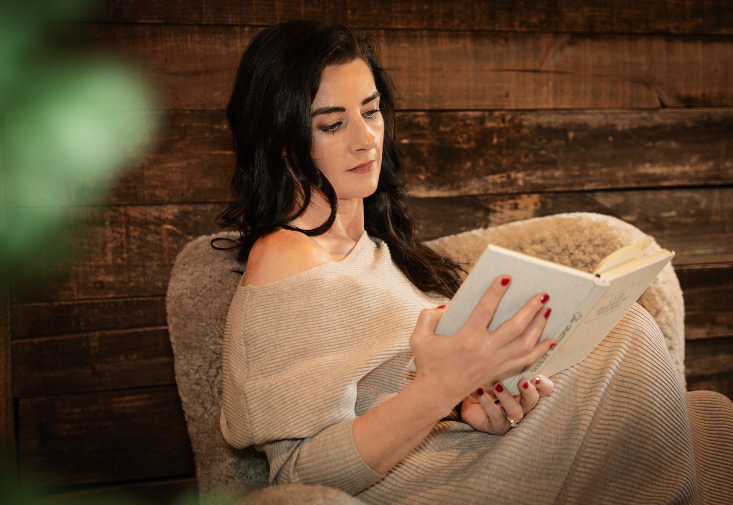 Young woman with red nails relaxing in cozy chair reading a book against rustic wooden wall background.