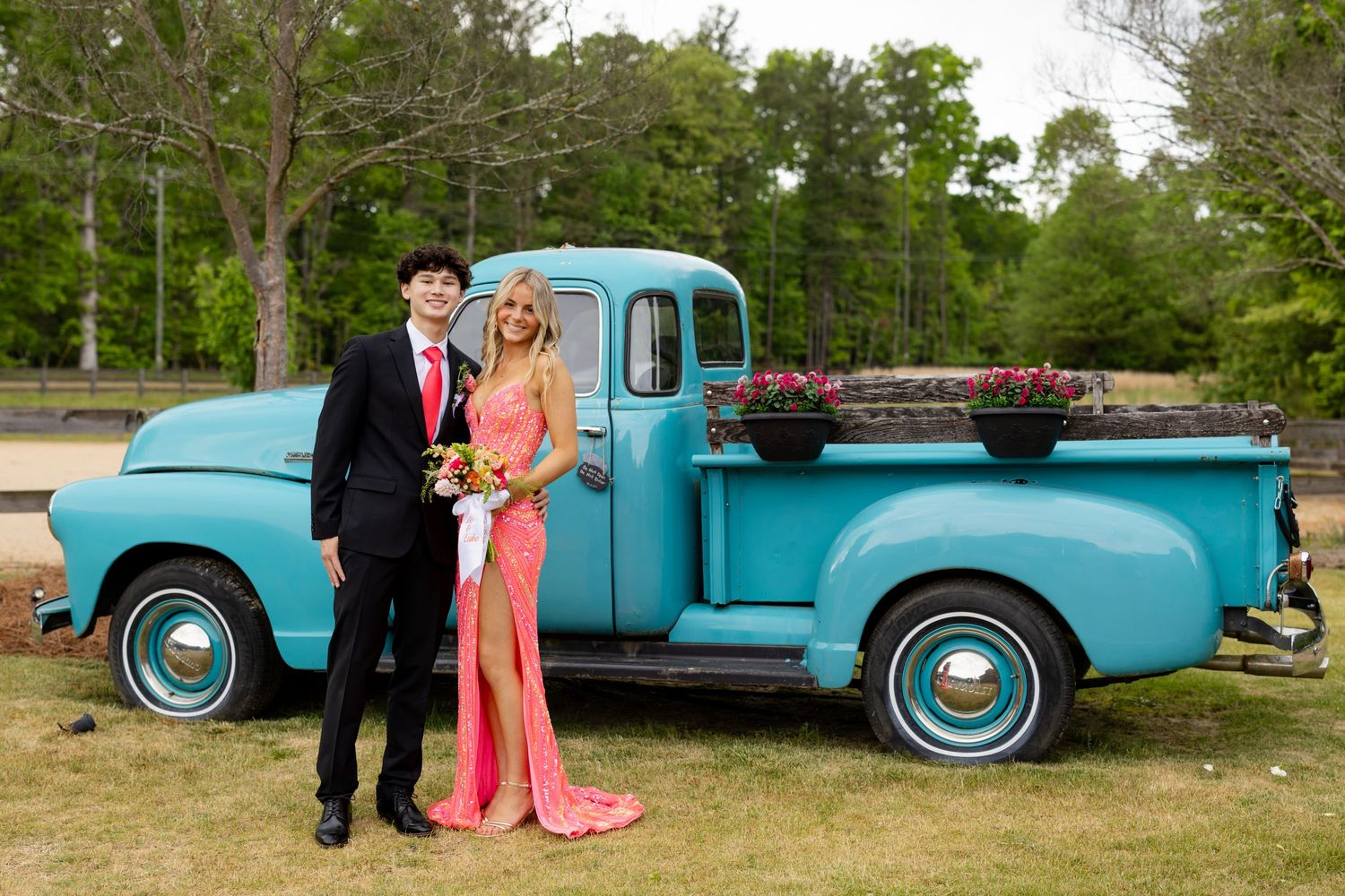 A couple in prom attire poses beside a vintage turquoise pickup truck decorated with flowers outdoors.