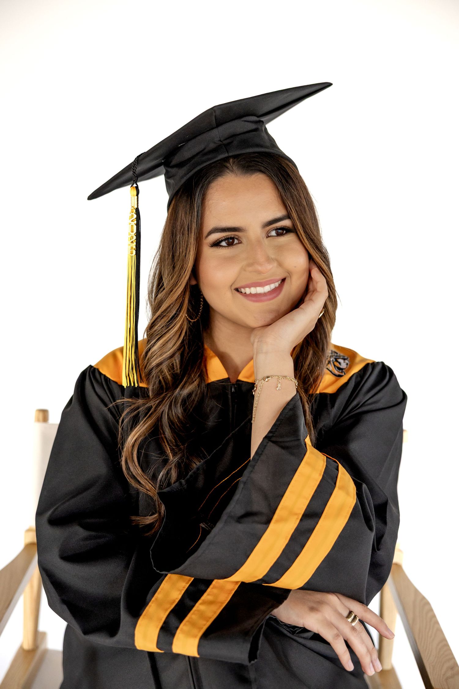 Smiling young woman in black and gold graduation cap and gown posing confidently for her graduation portrait.