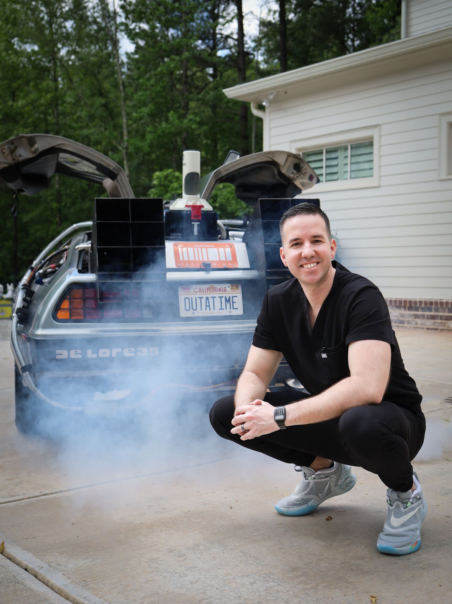 Man in black outfit crouching beside a DeLorean time machine replica with OUTATIME license plate and smoke effects.
