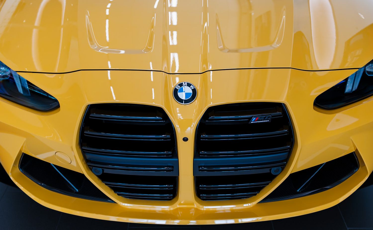 Yellow BMW M3 sports car front grille and hood detail showcasing iconic kidney grille design and blue roundel logo.