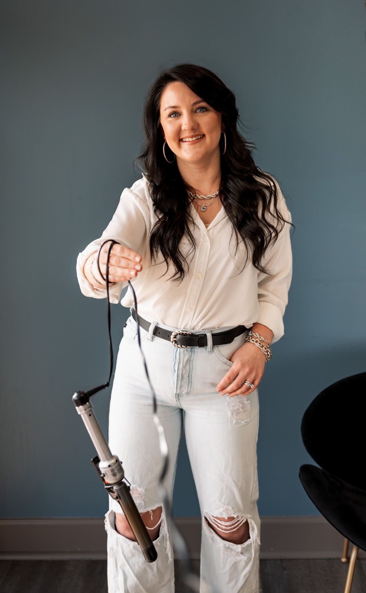 Woman in white blouse and ripped jeans holding a curling iron, smiling in a modern hair salon.