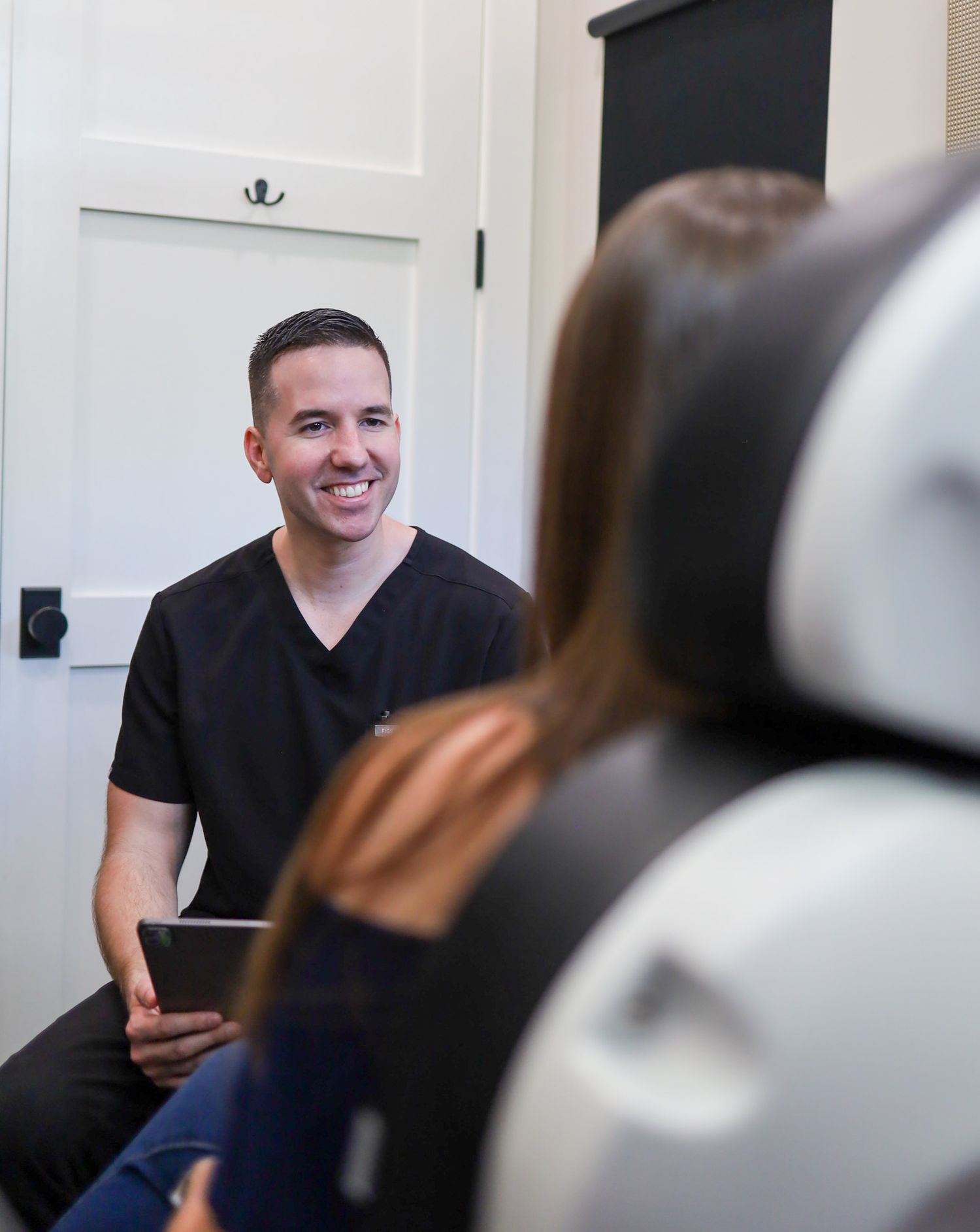 A smiling male dentist in black scrubs consults with a patient in a dental chair while holding a tablet.