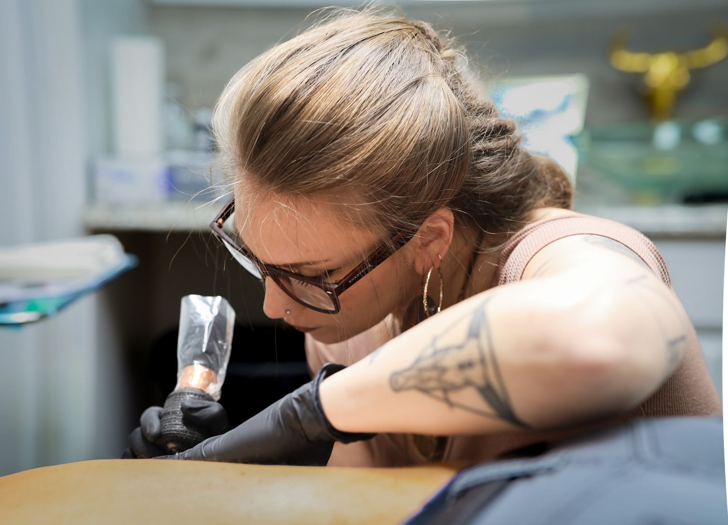Female tattoo artist with glasses and tattoos carefully inking a client's arm in a professional tattoo studio.