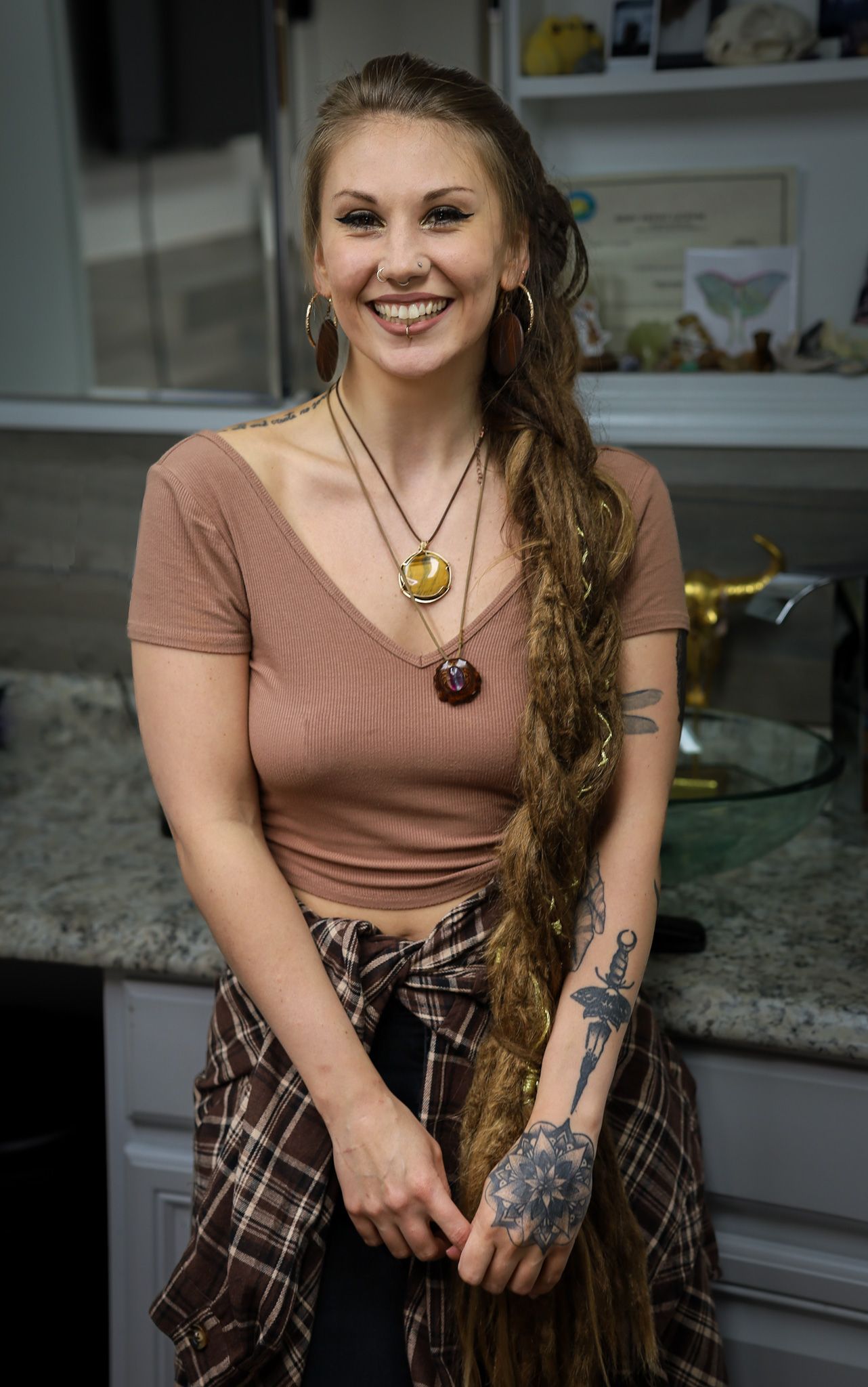 Smiling young woman with long wavy hair, tattoos, wearing a pink crop top and plaid shirt tied around her waist.