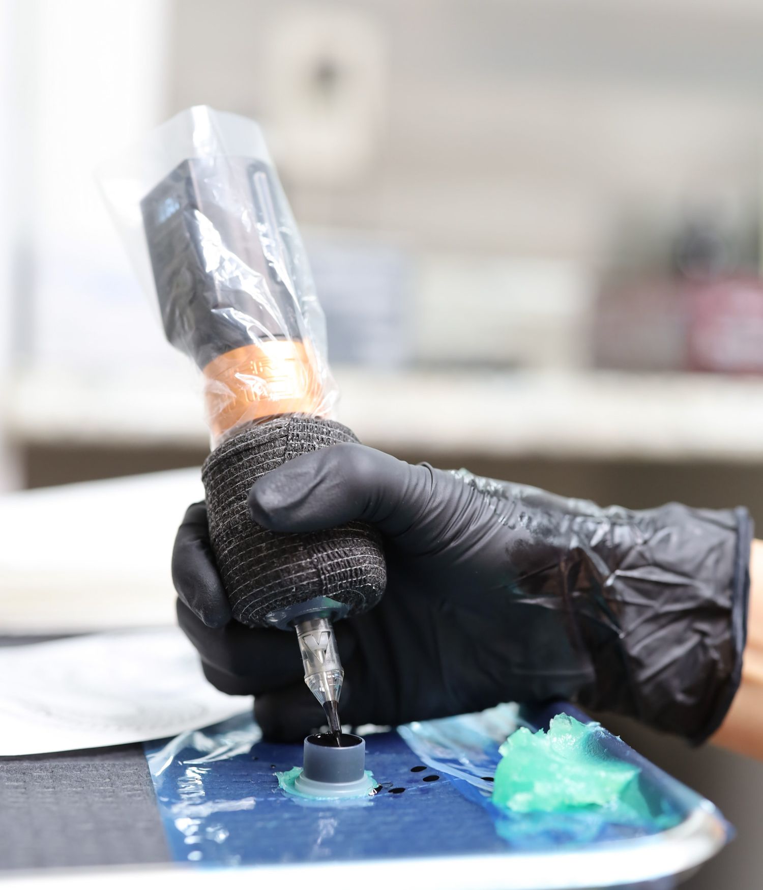 Tattoo artist wearing black gloves holds a wrapped tattoo machine over an ink palette with green pigment.