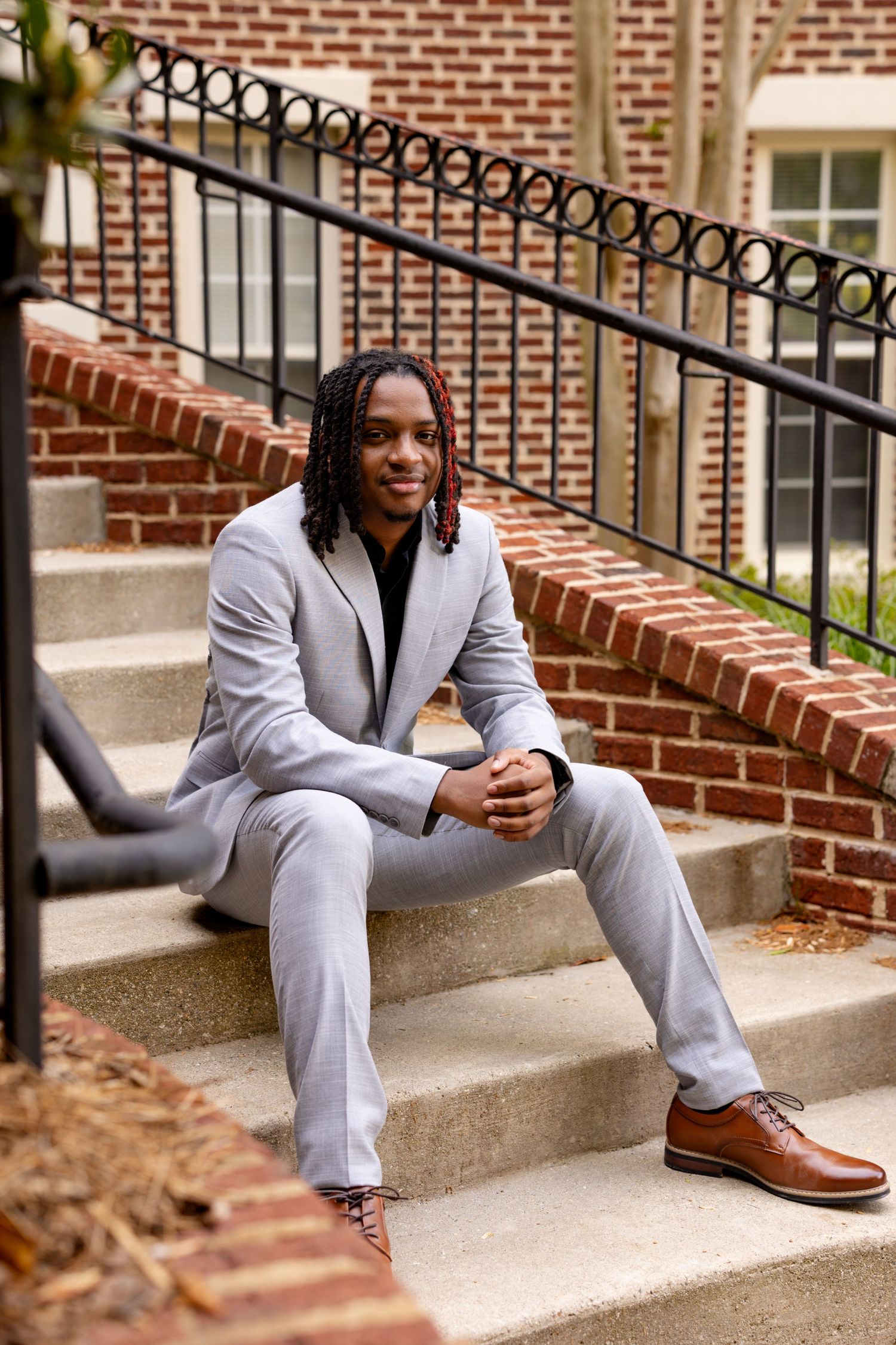 Young man in gray suit with braids sits smiling on brick building steps, wearing brown oxford shoes.